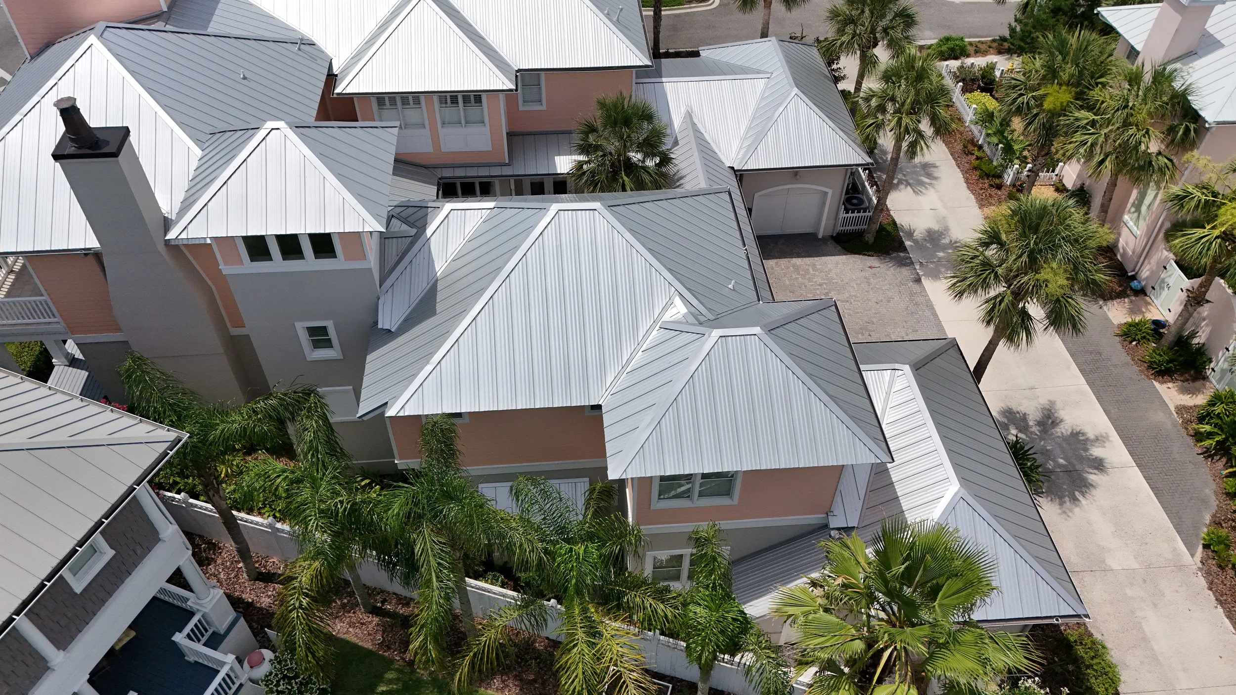 Aerial view of a neighborhood with pastel-colored houses, metal roofs, palm trees, and concrete sidewalks.
