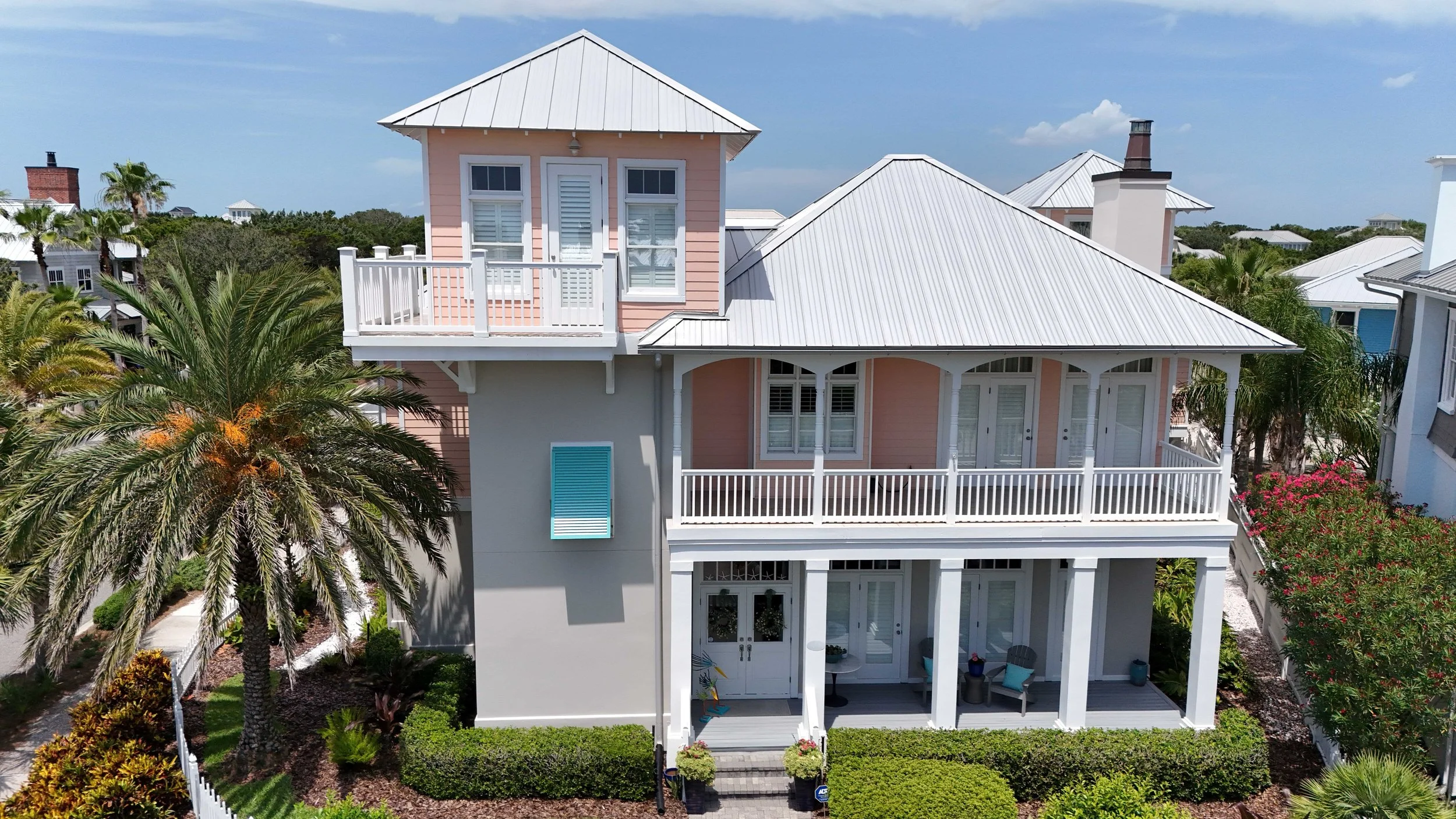 A multi-story beach house painted in pastel pink with white trim, featuring a front porch, balcony, and metal roof, surrounded by tropical plants and palm trees in a coastal neighborhood.