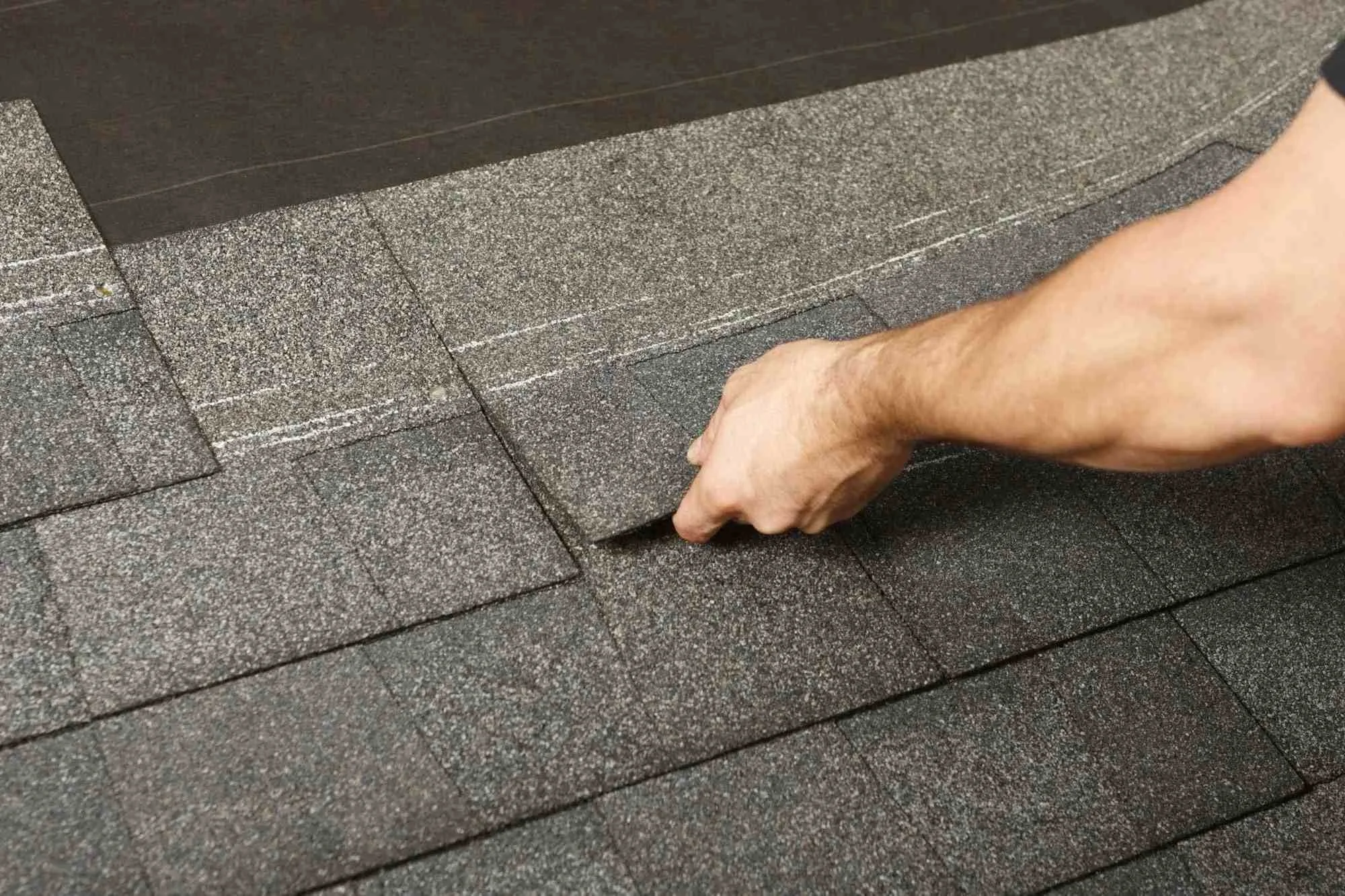 A person installing asphalt shingles on a roof, with a hand holding a shingle and laying it down over other shingles.