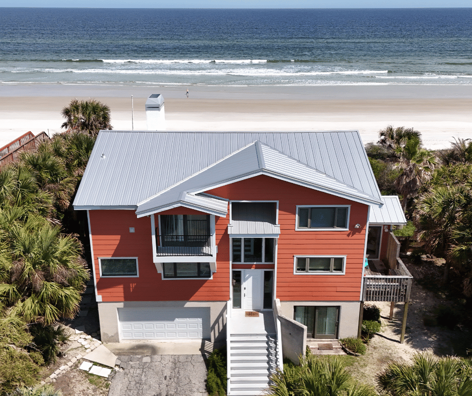 An aerial view of a three-story house painted in red with white trim, located near a sandy beach and ocean, surrounded by palm trees.