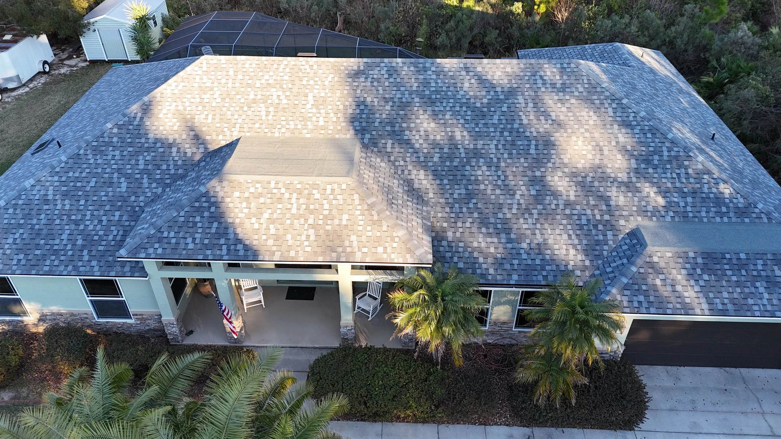 Aerial view of a house with a tiled roof, front porch with white chairs, palm trees, bushes, and a concrete driveway.