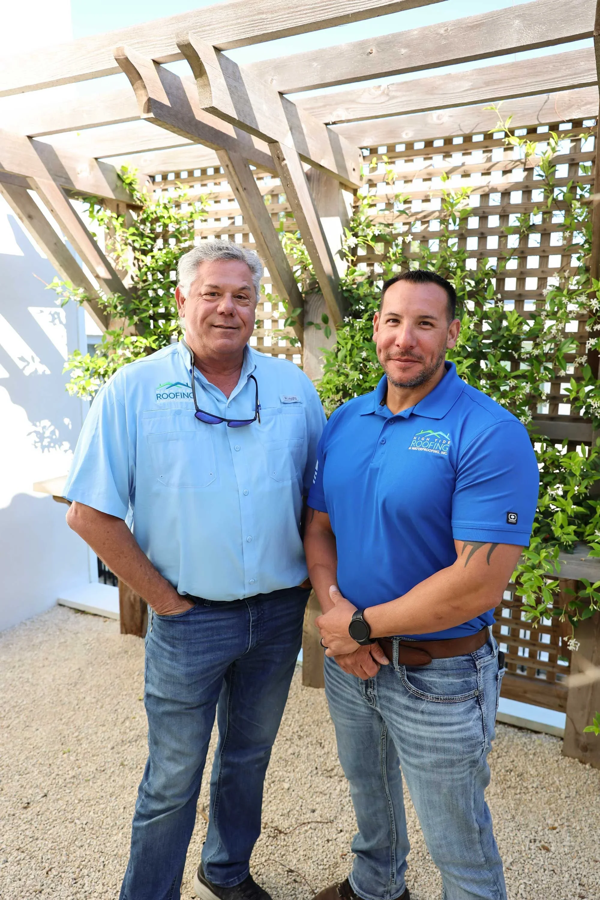 Two men standing outdoors in front of a wooden trellis and greenery, wearing branded shirts with company logo, smiling and posing for the photo.