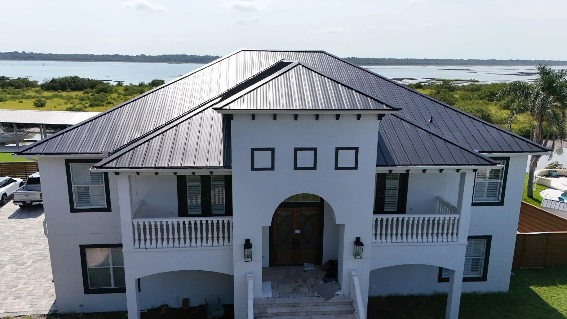 A white two-story house with black trim and a metal roof, situated near a body of water with greenery and trees in the background.