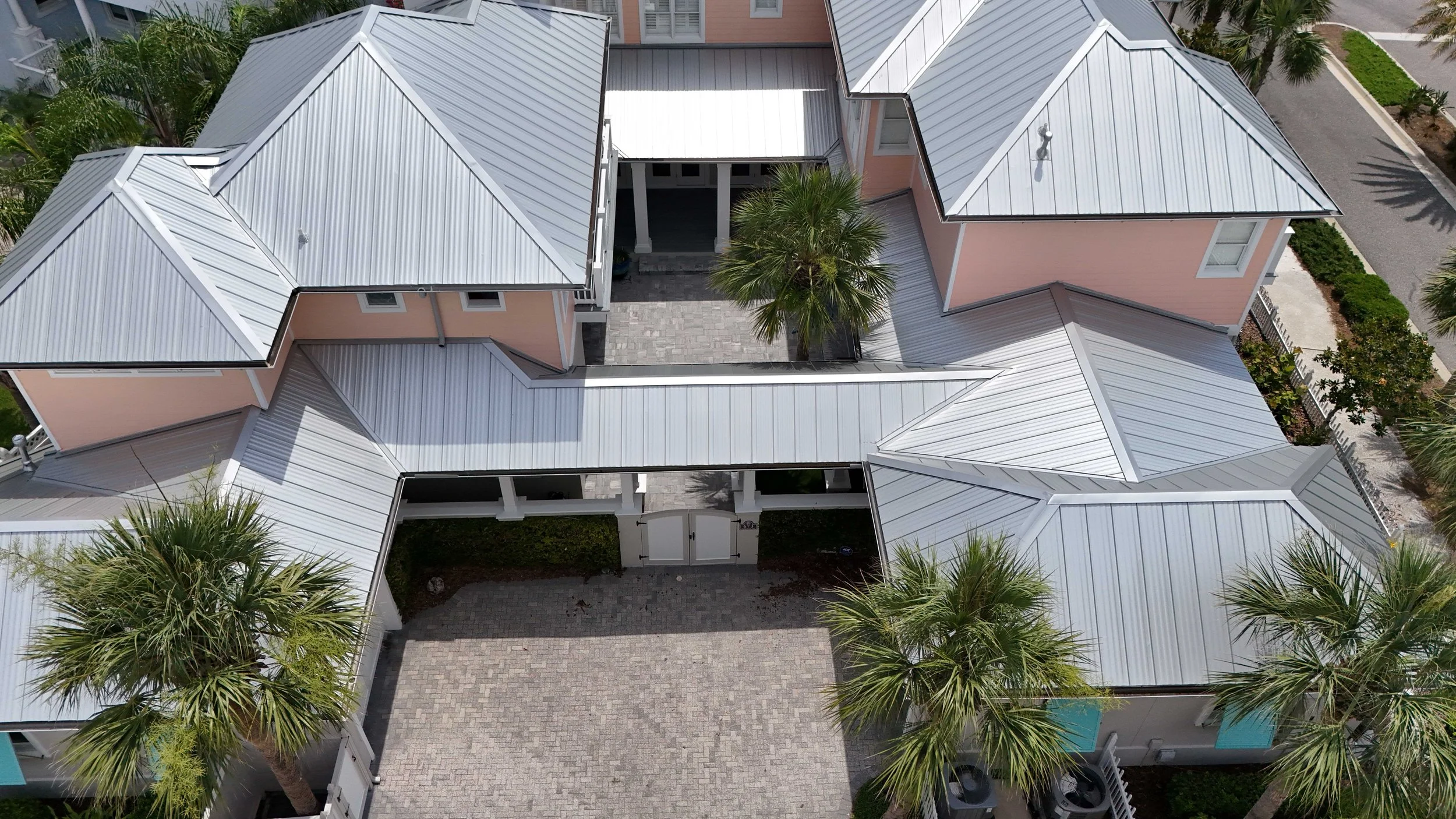 Aerial view of a residential complex with interconnected buildings featuring metal roofs, a central courtyard with a tree, palm trees surrounding the area, and parking spaces on the side.