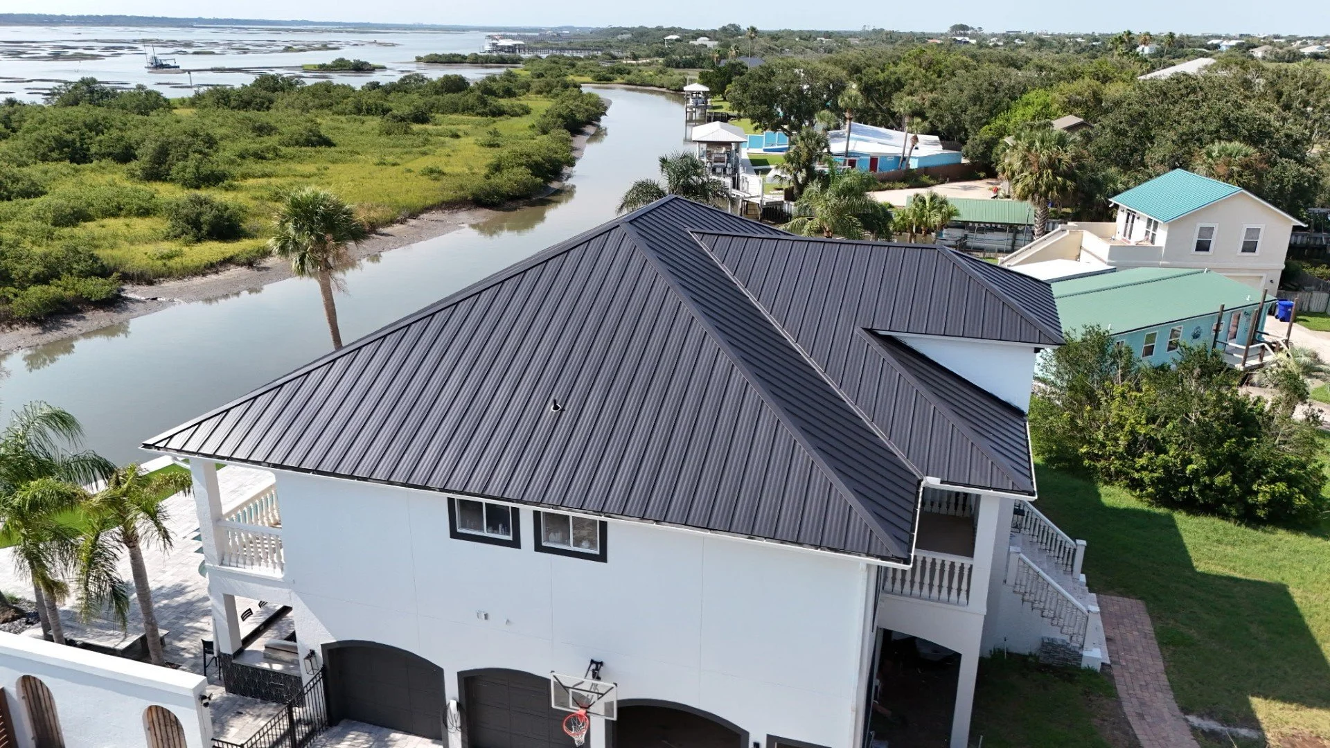 Aerial view of a modern two-story house with a dark metal roof, situated near a waterway with lush greenery. The house has a basketball hoop on the driveway, a small outdoor patio, and a backyard with trees and a brick pathway. Other colorful houses 