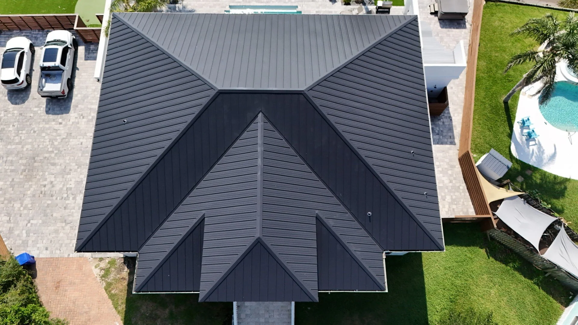 Aerial view of a house with a dark gray metal roof, a driveway with parked cars, a backyard with a swimming pool, lounge chairs, shade sails, and a well-maintained lawn.