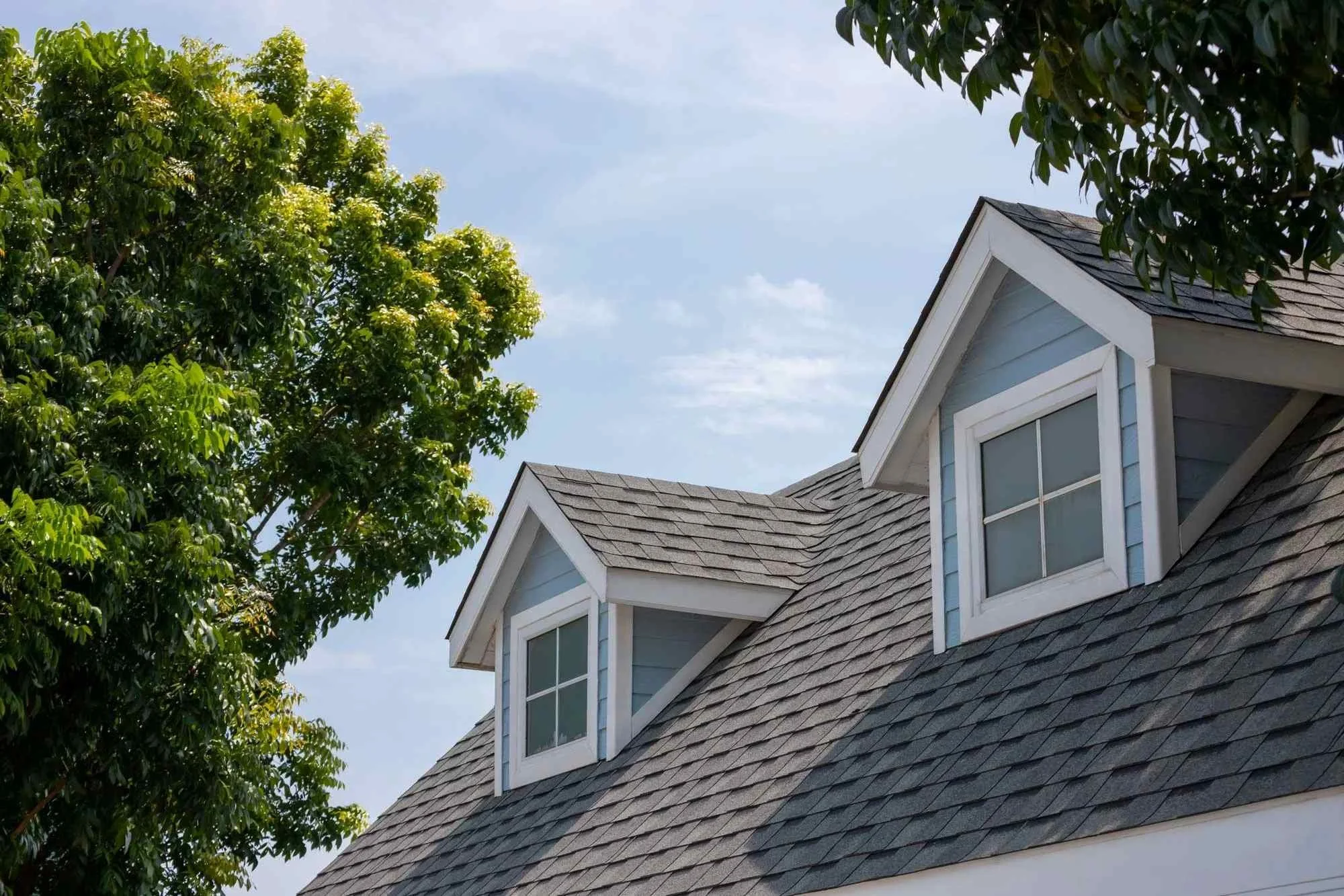 View of a gray-shingled house with dormer windows and a tree with lush green leaves against a blue sky with some clouds.