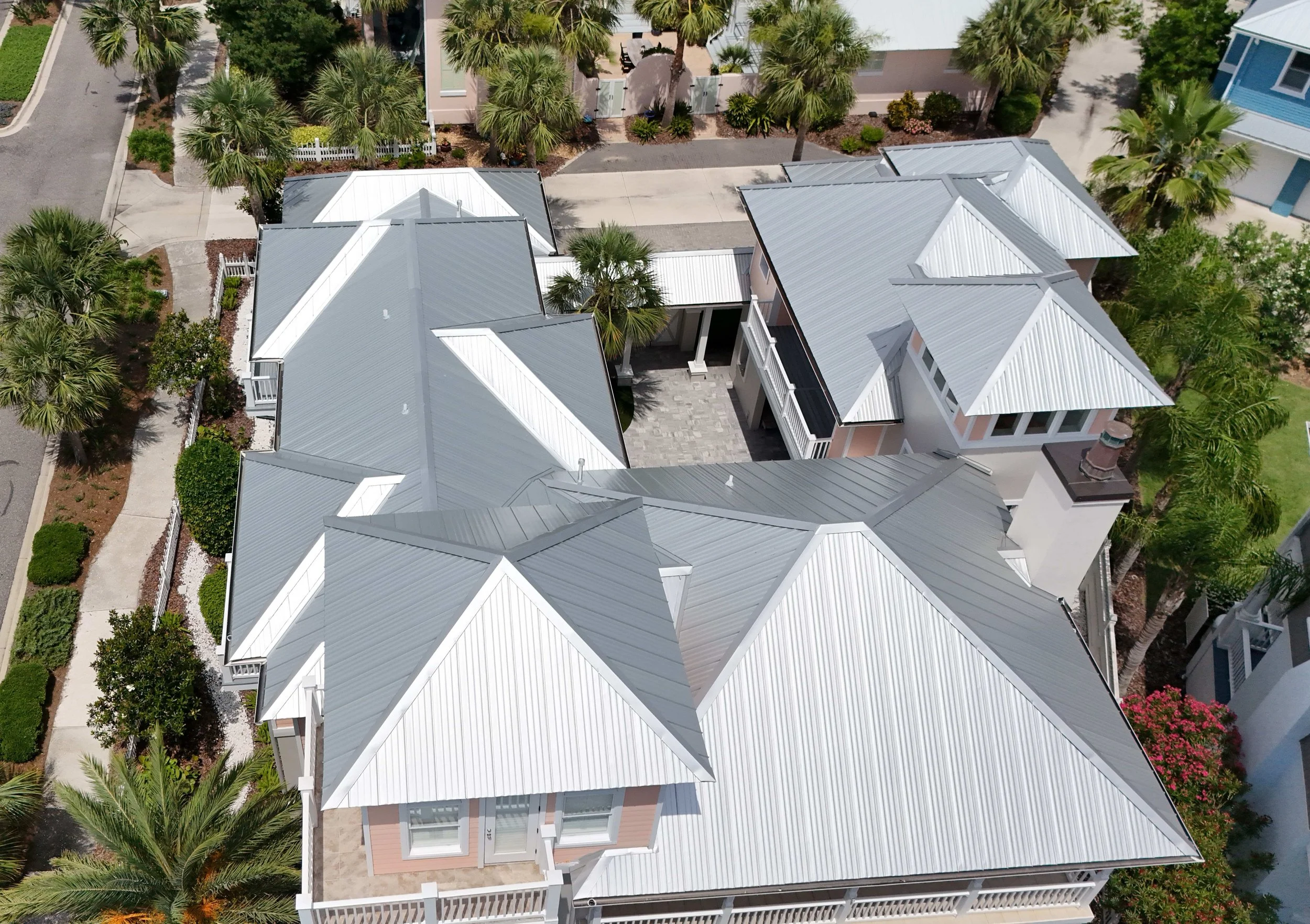 Aerial view of houses with gray metal roofs, surrounded by palm trees and greenery, in a suburban neighborhood.