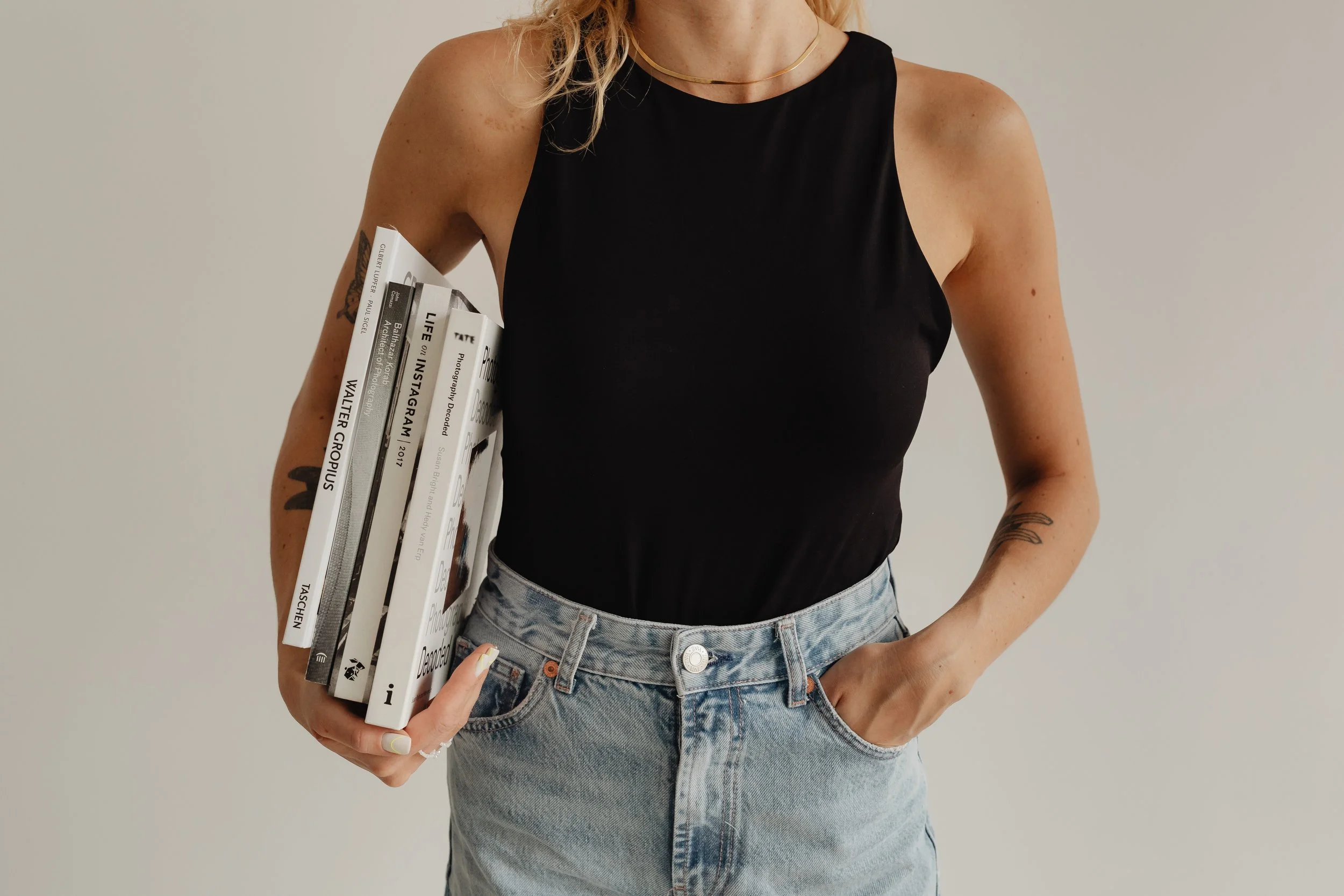 Woman in a black sleeveless top and light blue jeans holding books against a plain background.