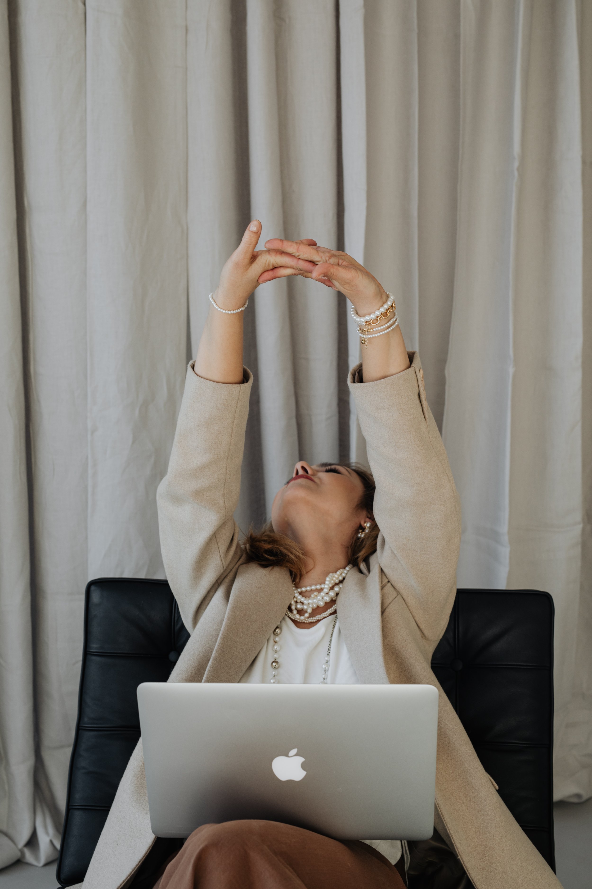 A woman wearing pearl jewelry sits at a desk holding an iPad above her head with both hands. She is in front of a laptop with an Apple logo, seated in a black chair with a beige blazer, white top, and brown pants. Behind her are cream-colored curtains.