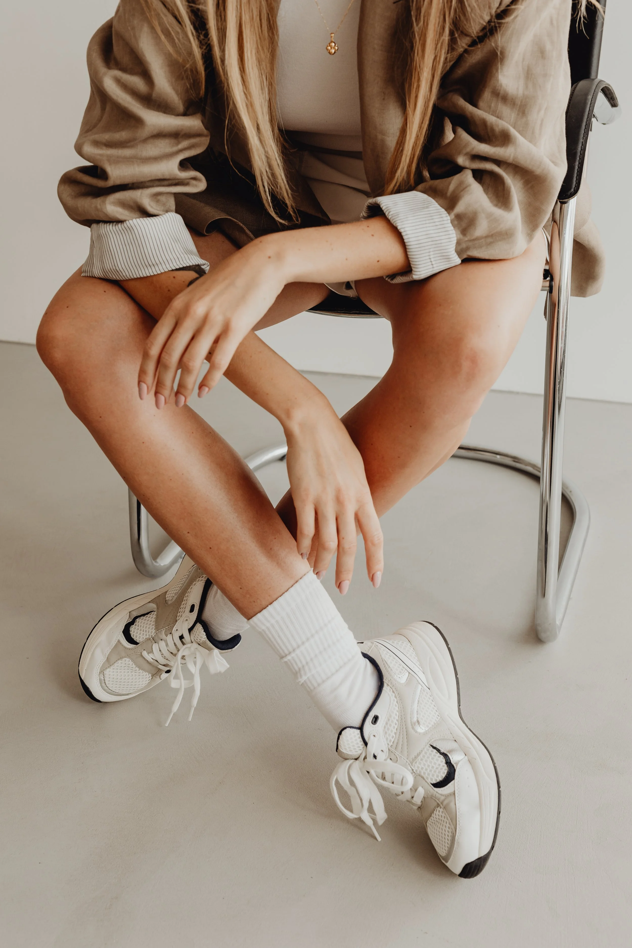 A person sitting on a chair wearing white sneakers, white socks, beige shorts, a beige blazer, and a white top, with long blonde hair and a gold necklace, with their legs crossed and hands resting on their knees.