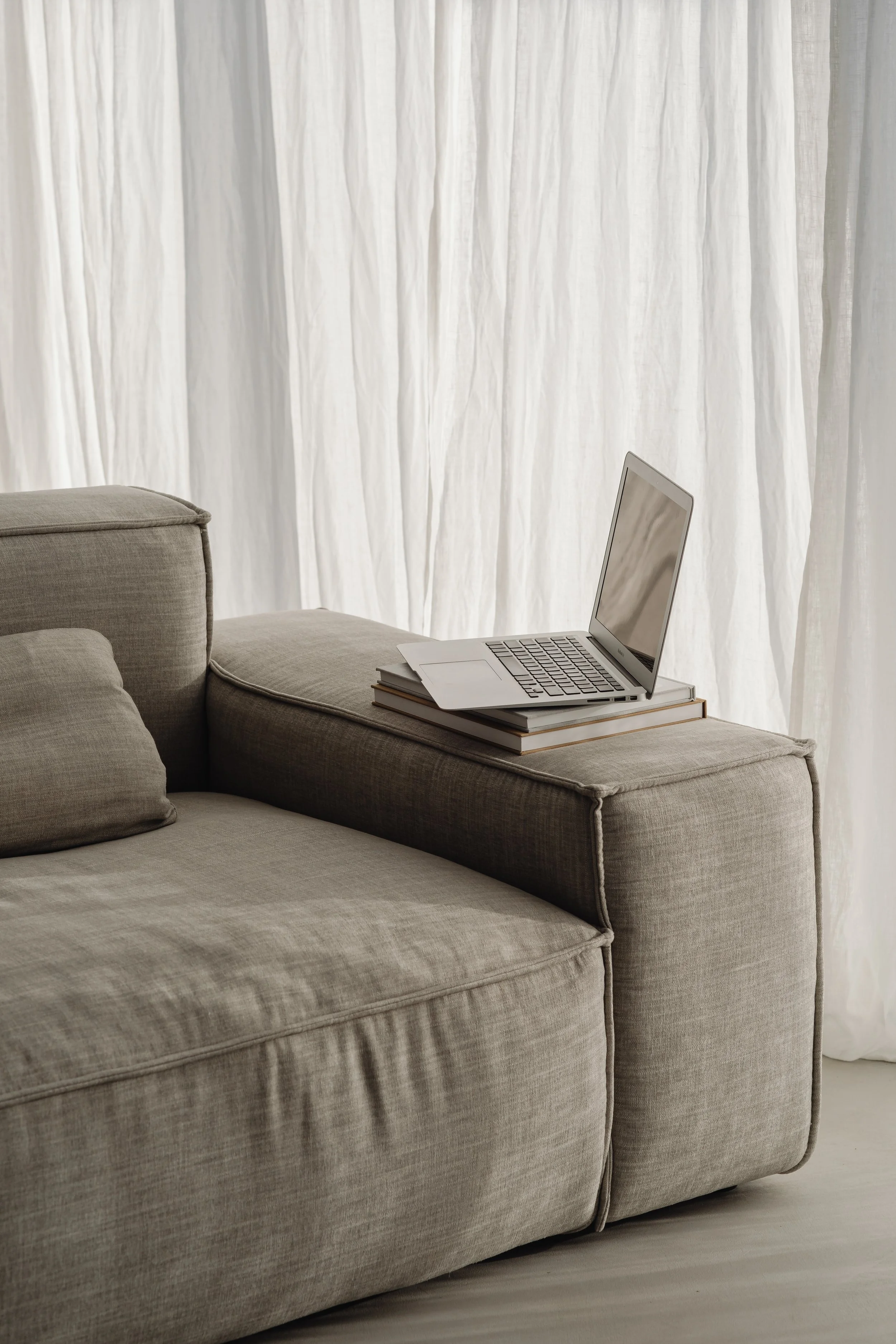 A beige fabric sofa with cushions, a three-layered book, and an open silver laptop on the armrest, with a white curtain in the background.
