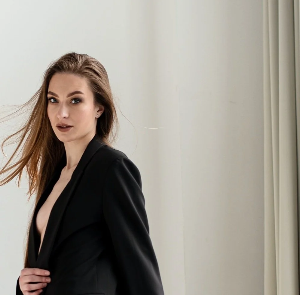 A woman with long brown hair wearing a black blazer and makeup, standing in front of a white wall and a window with beige curtains.