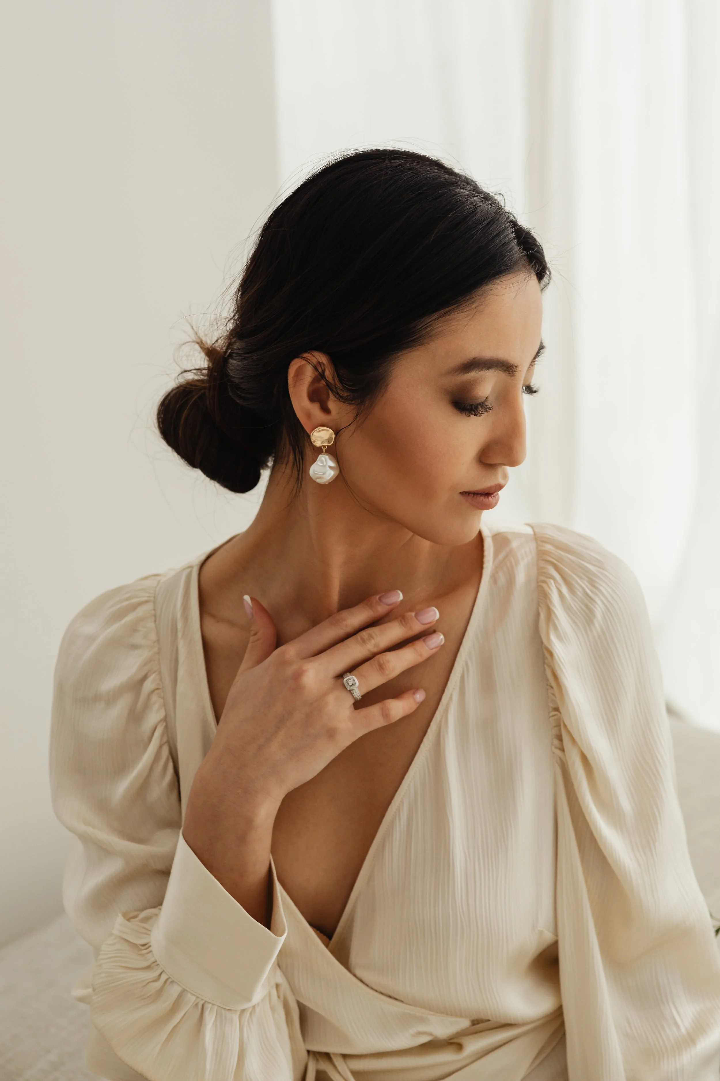 A woman with dark hair styled in a low bun wearing pearl and gold earrings, a diamond ring, and a cream-colored, long-sleeved blouse, sitting with her eyes closed and her hand resting on her chest, in a softly lit room.