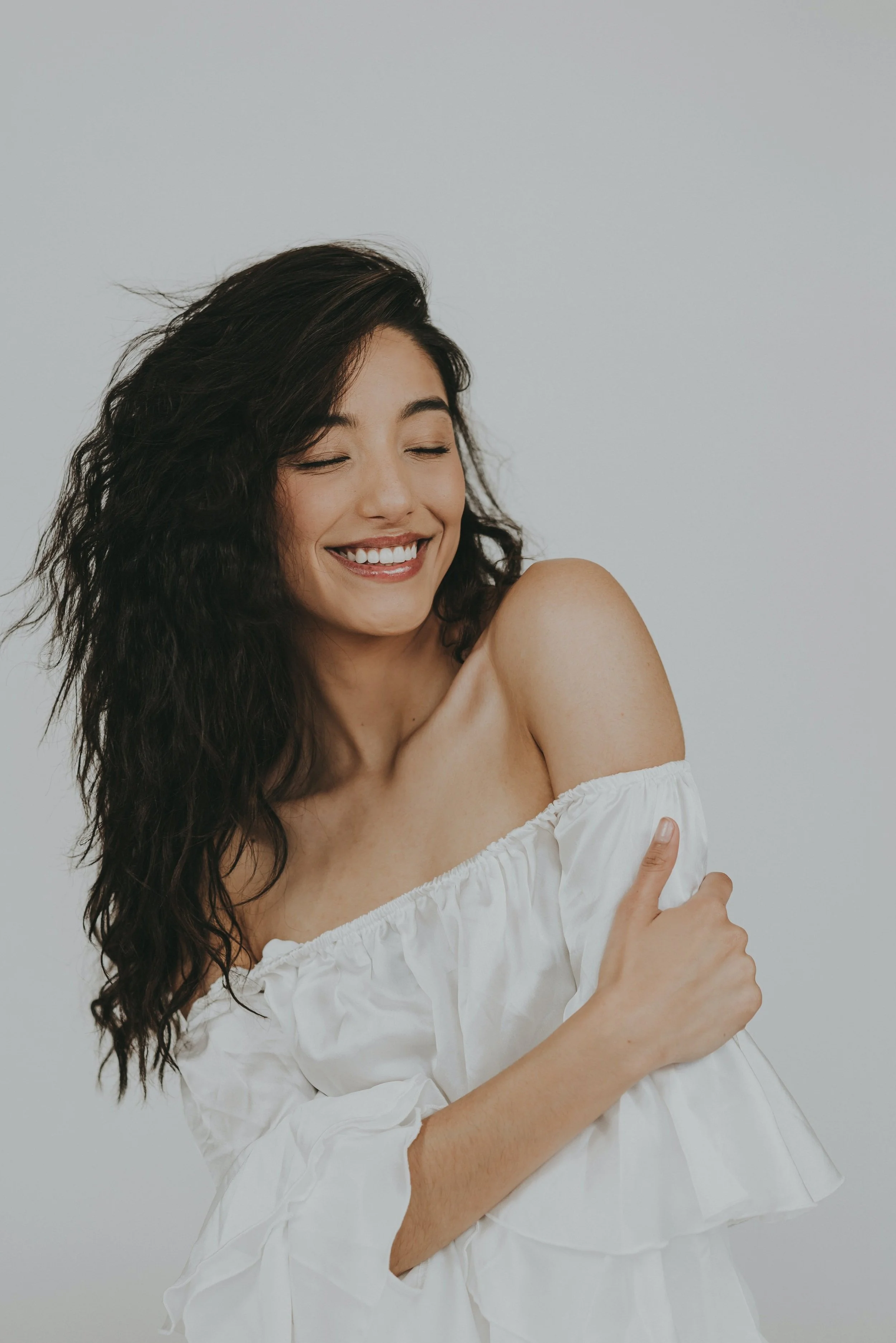 Smiling woman with curly dark hair, wearing an off-the-shoulder white dress, posing against a plain light gray background.