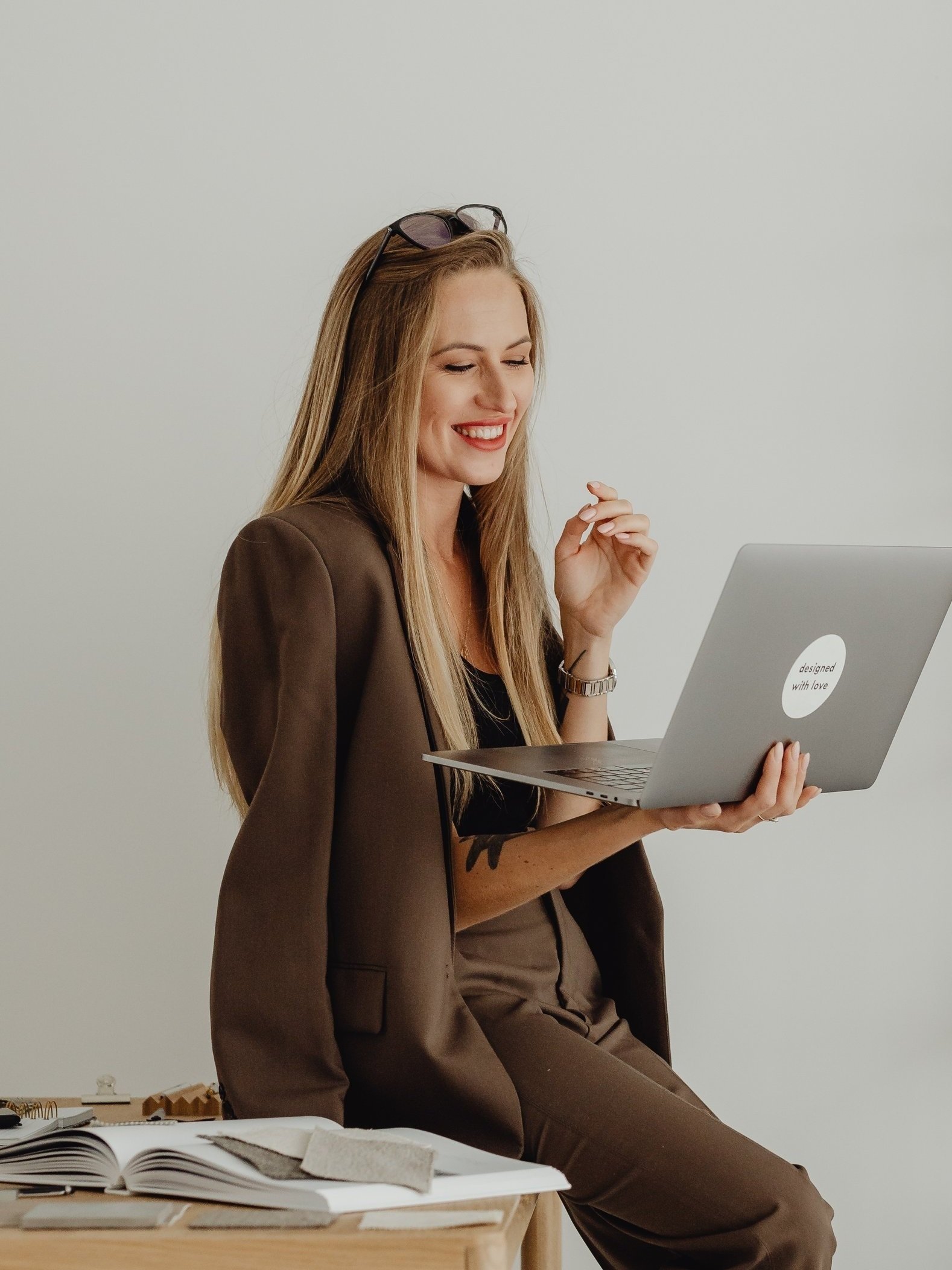 A woman in brown suit sitting on a table, smiling and holding a laptop with a sticker that reads "designed with love." She has sunglasses on her head and is in a workspace with sketchbooks and drawings on the table, against a plain white wall.
