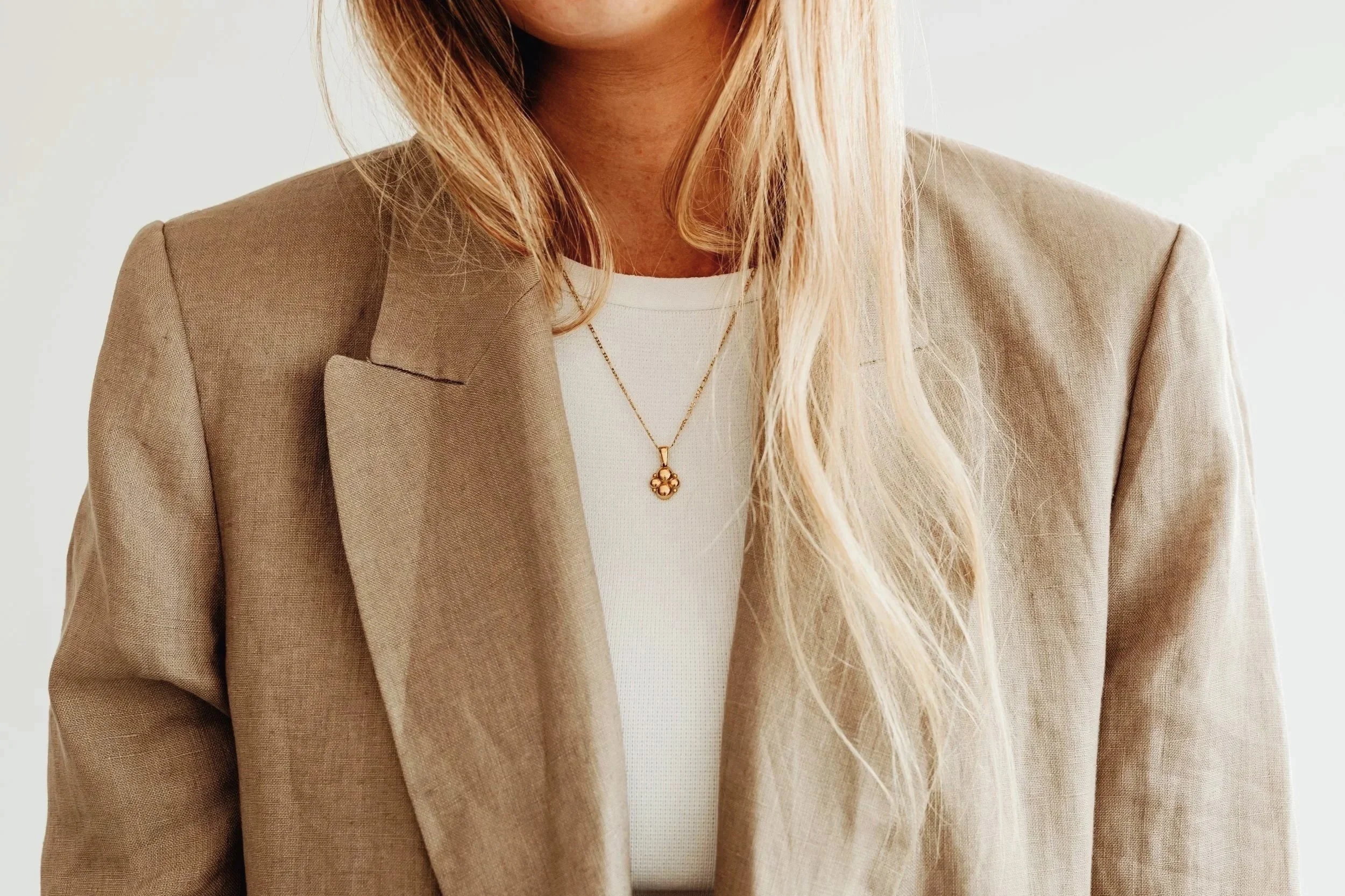 Close-up of a woman wearing a beige blazer, white top, and a gold pendant necklace, with long blonde hair.