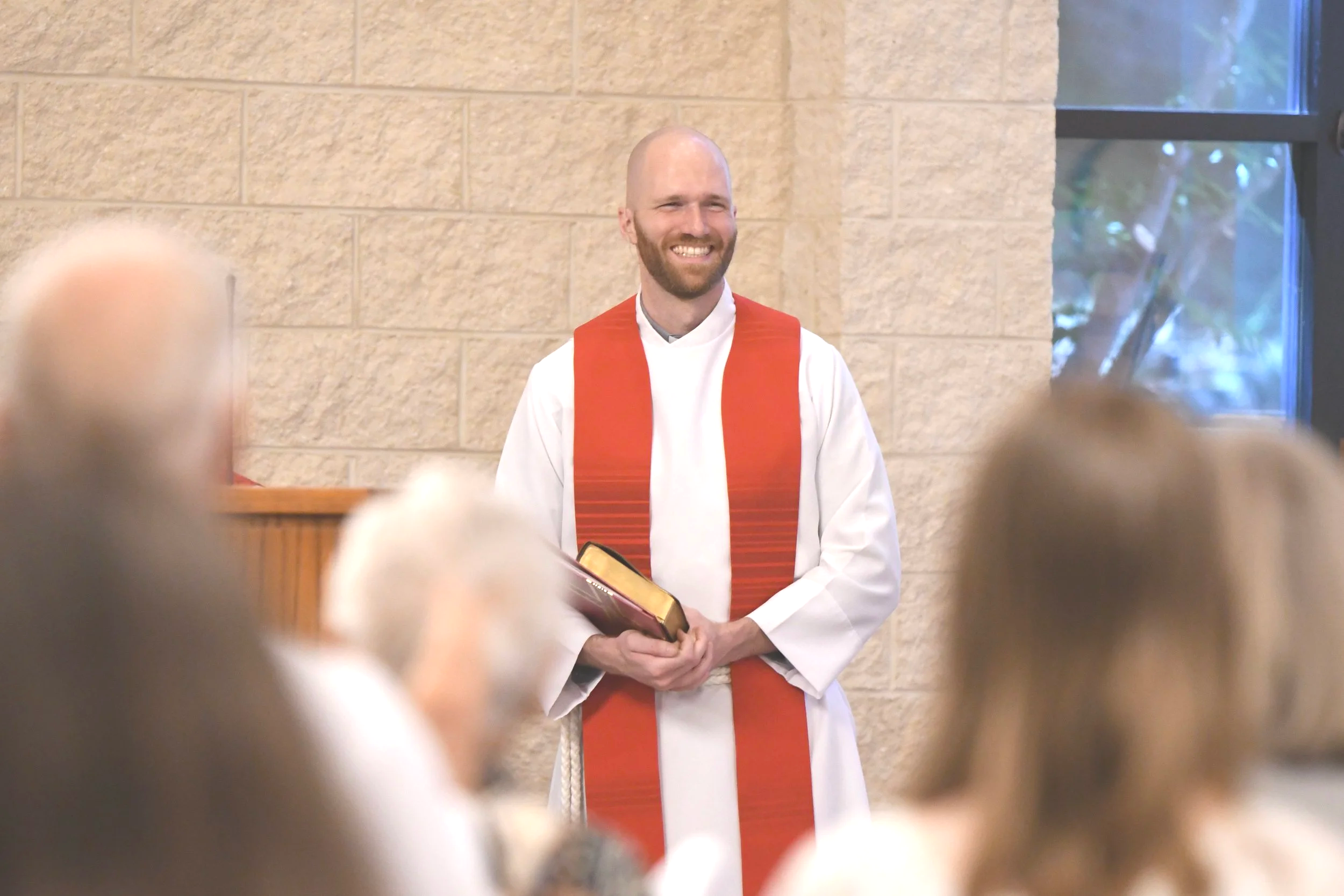 A smiling man with a beard wearing a white robe and an orange stole, inside a building with tan brick walls and a window, during a religious ceremony, holding a book.
