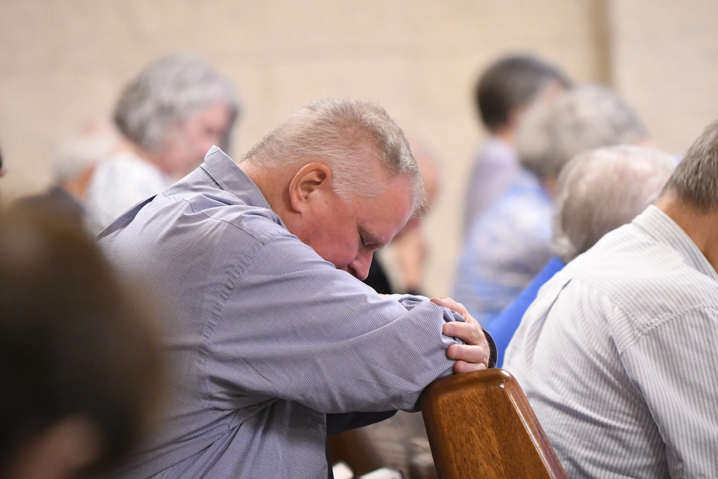 A man with short hair and a checkered shirt bows his head and prays with his eyes closed at a gathering, sitting at a wooden table.