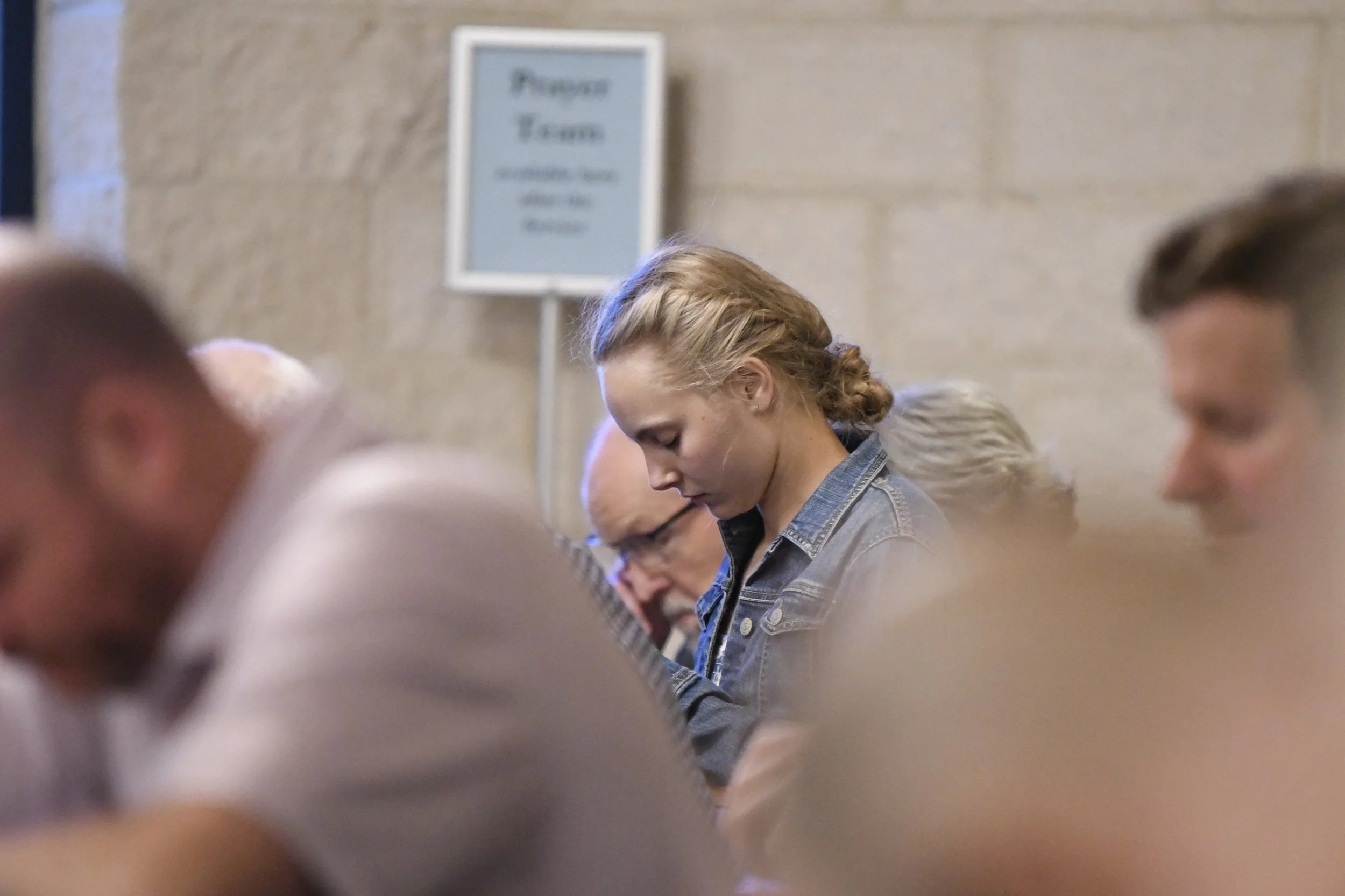 People sitting and standing in a room, with a woman with red hair and a denim jacket prominent in the center, and a blurred foreground with people's heads and hair.