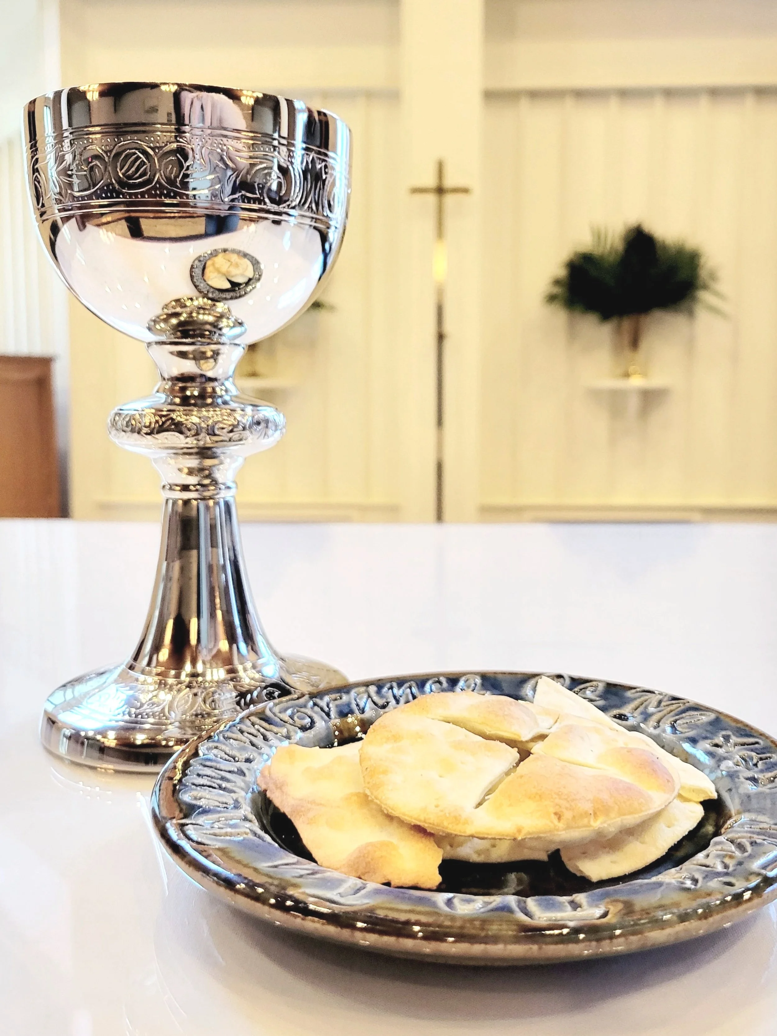 Silver ornate chalice with intricate detailing, reflecting the surrounding environment, and a plate with bread slices on a white surface in a well-lit room with cream-colored walls, a cross, and a plant in the background.