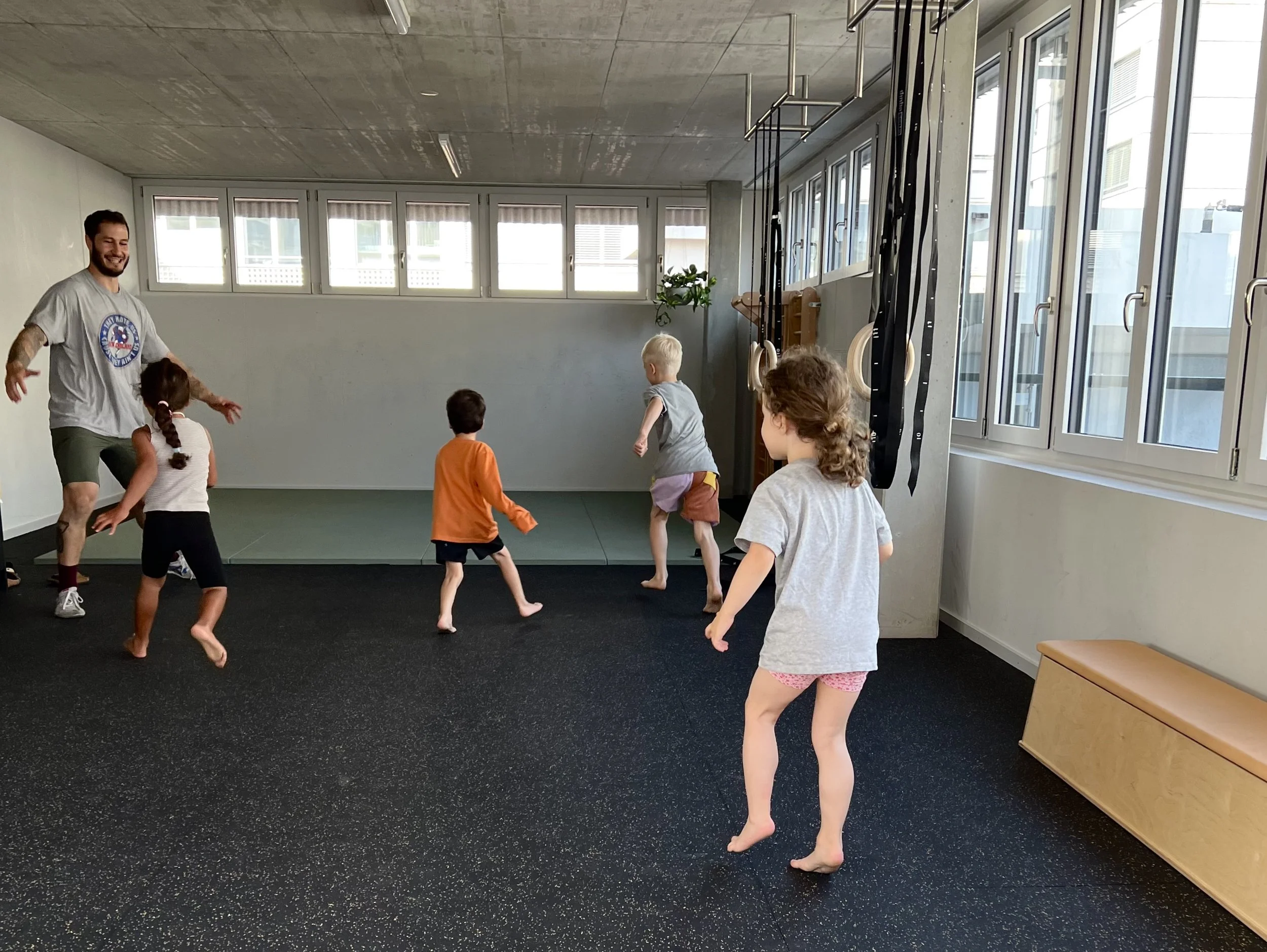 Ein Trainer führt Kinder in einem Fitnessraum beim Sport. Die Kinder springen und bewegen sich auf einem schwarzen Boden mit kleinen bunten Punkten. Im Hintergrund sind Gymnastikmatten, Turnrings und Seile an der Wand befestigt. Große Fenster lassen viel Tageslicht in den Raum.
