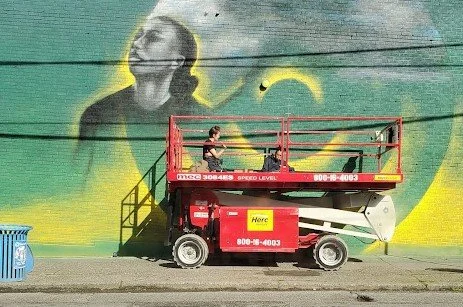 Seattle Storm mural in progress, photographer unknown. Image description: A painter perches on a red scissor lift in front of a building covered by a mural. The mural is unfinished and currently features a green and yellow background with a Storm pla
