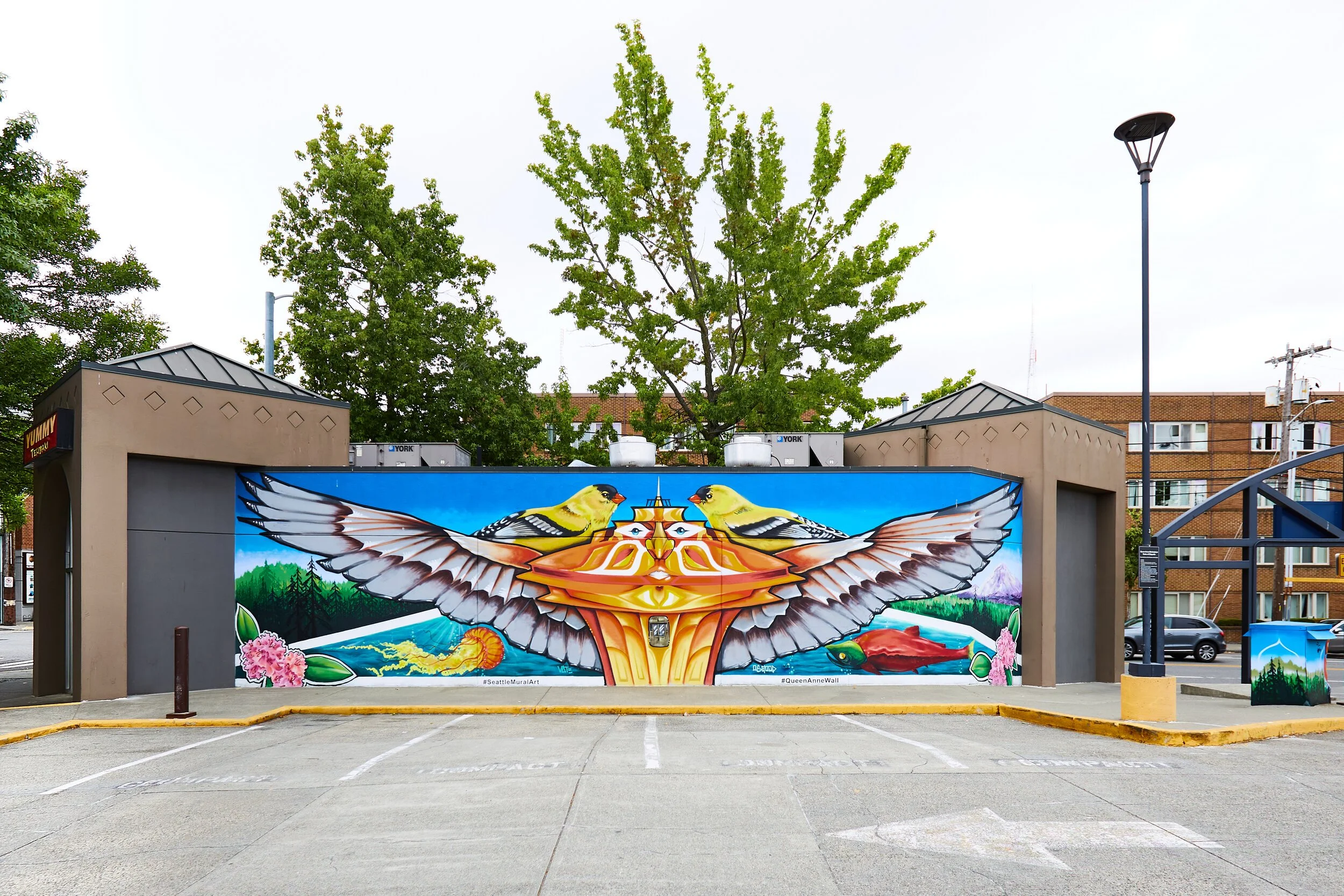 Queen Anne Marketplace mural, photo by UACC volunteer. Image description: In a small parking lot, the side of a building is covered in a large, wide mural. The mural features two yellow birds perched on the top of the Space Needle, with what appear t
