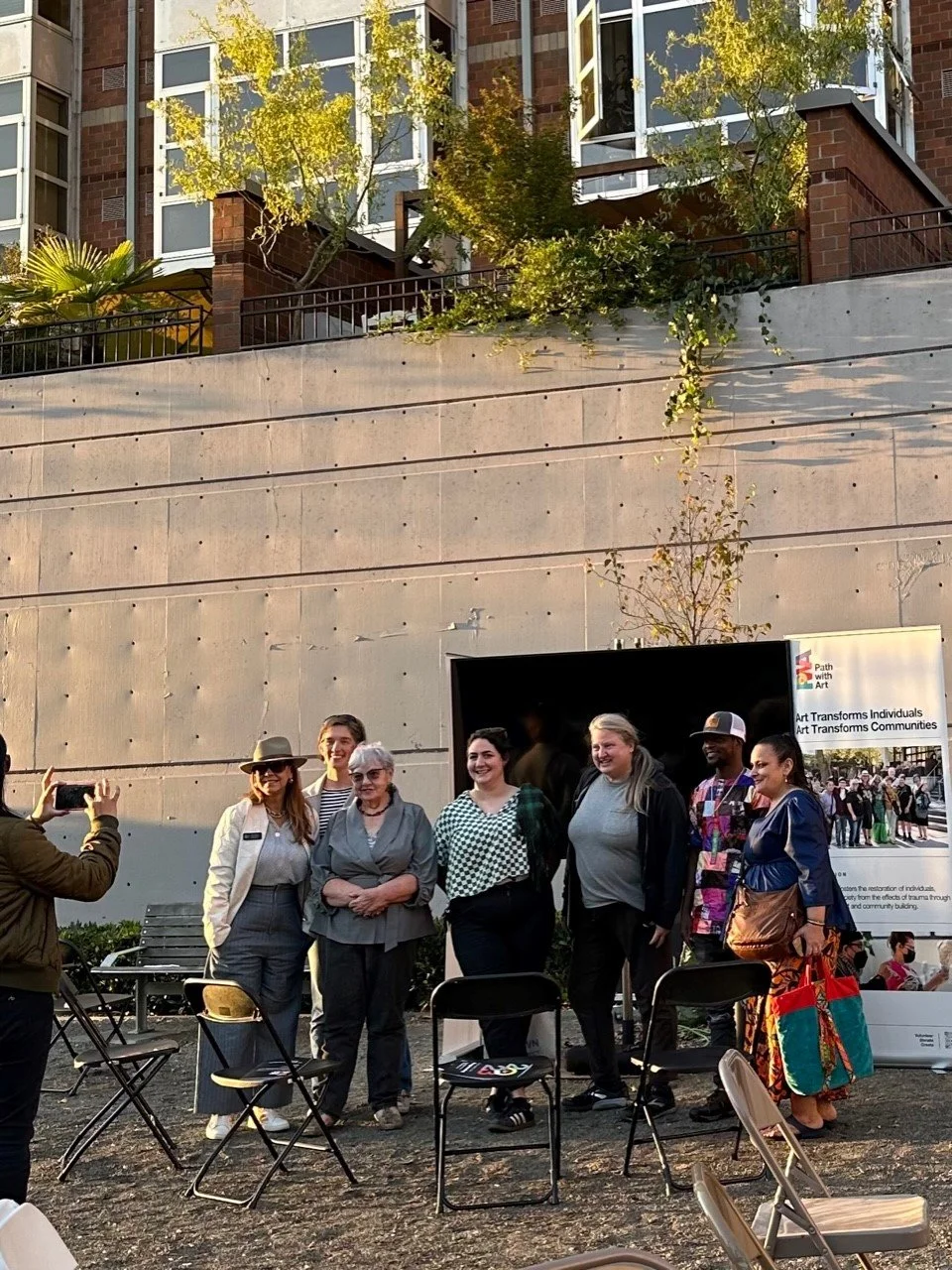 Path with Art group photo at Uptown Artists Reception, photo by UACC volunteer. Image description: 7 people stand in front of a monitor and the Path With Art banner. They are in Counterbalance Park, with golden hour light shining in from the west.