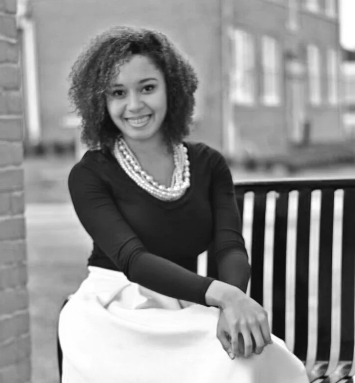 A woman with curly hair wearing a black top, a multiple-strand pearl necklace, and a white skirt, sitting on a bench outdoors with a smile.