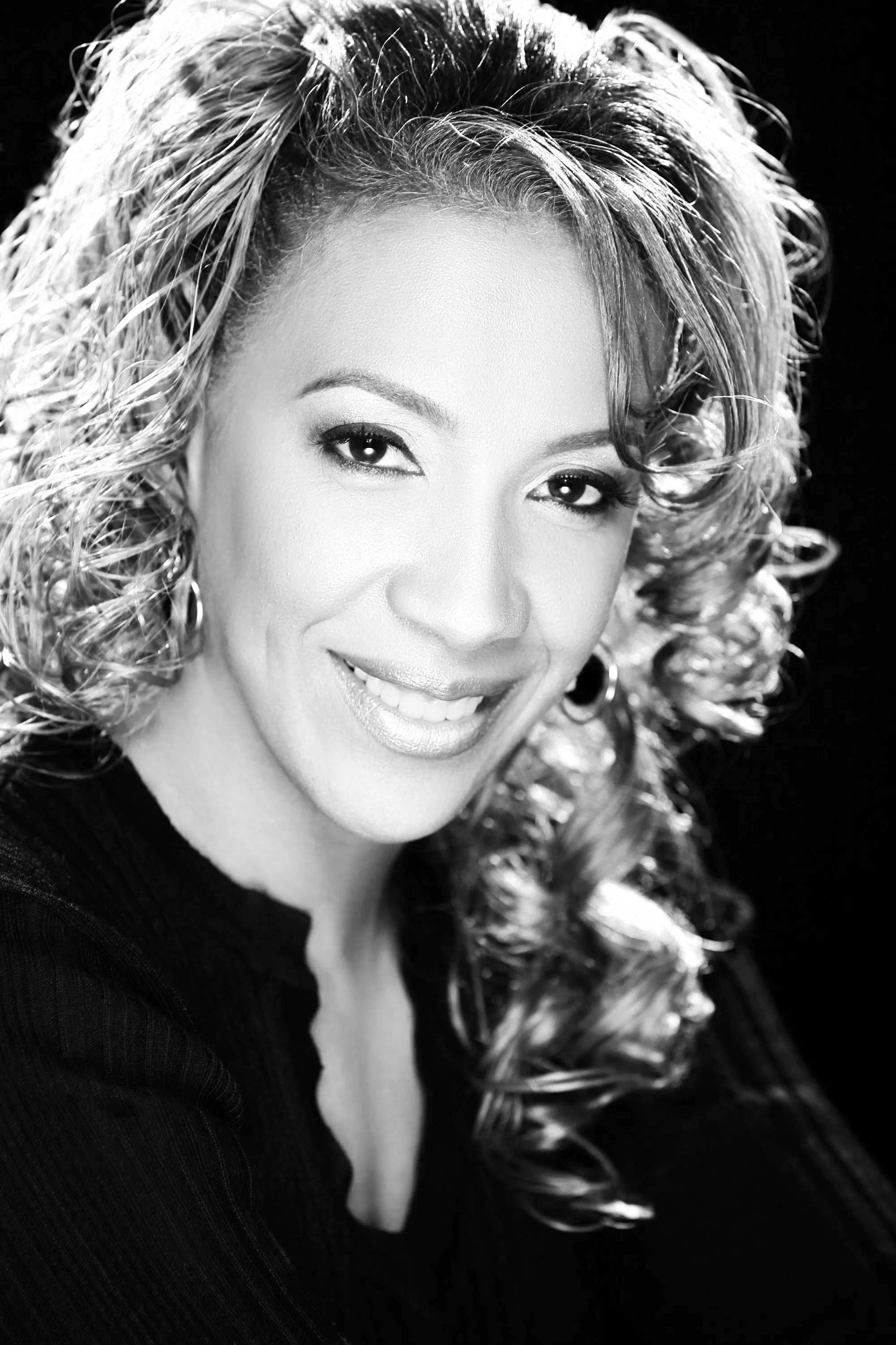 Black and white portrait of a woman with curly hair, smiling, wearing earrings and a dark top.