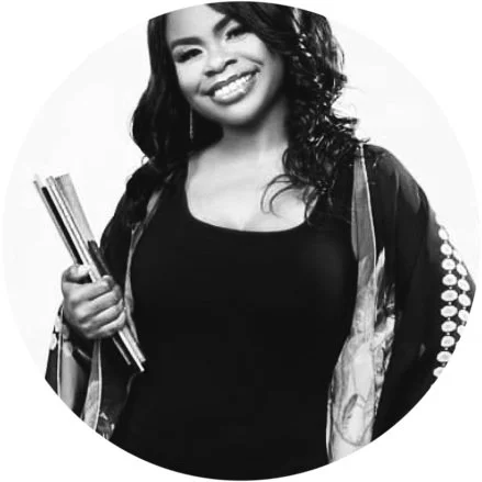 Young woman smiling, holding books or notebooks, wearing a dark top and a patterned jacket, with curly hair.