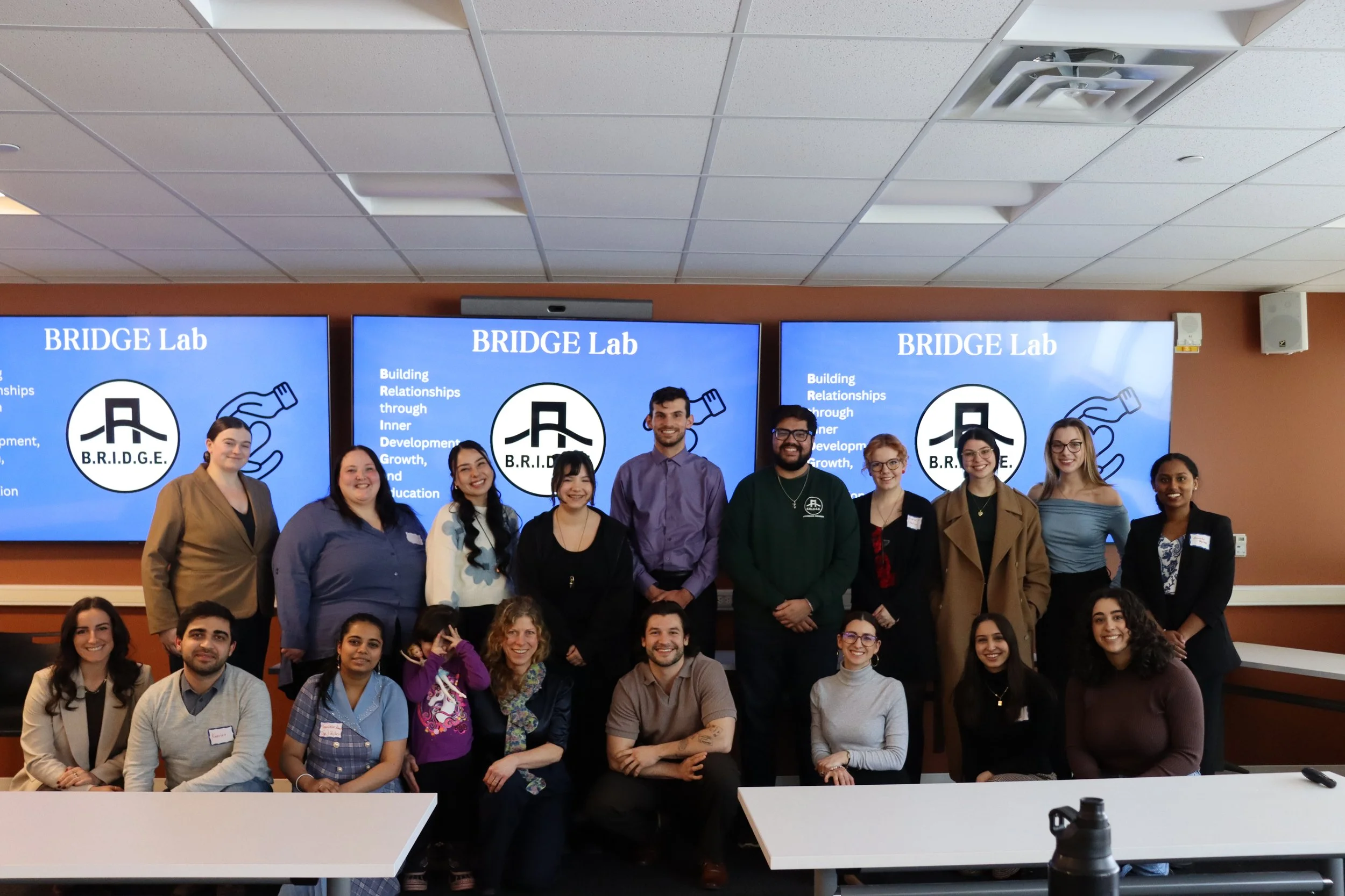 Group of diverse people posing for a photo in front of a screen with the text 'BRIDGE Lab' and a logo, inside a conference room.