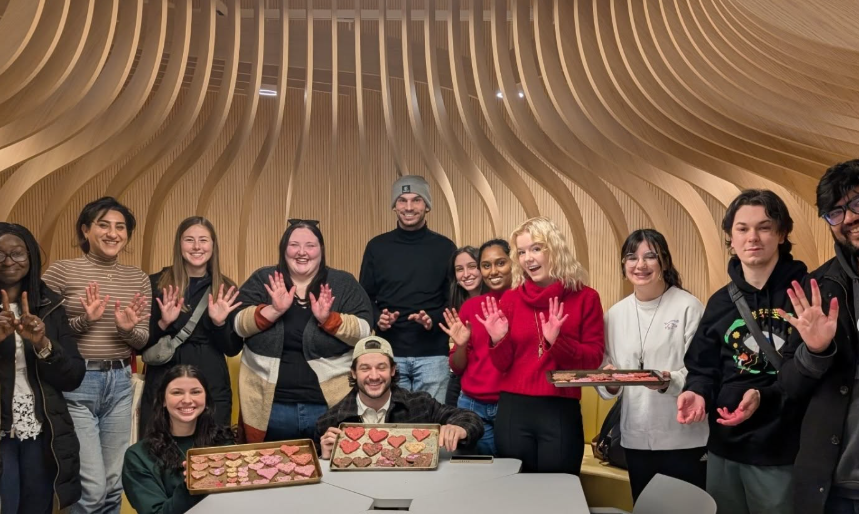 Group of people smiling, raising hands, with cookies on trays, in a modern wooden interior.
