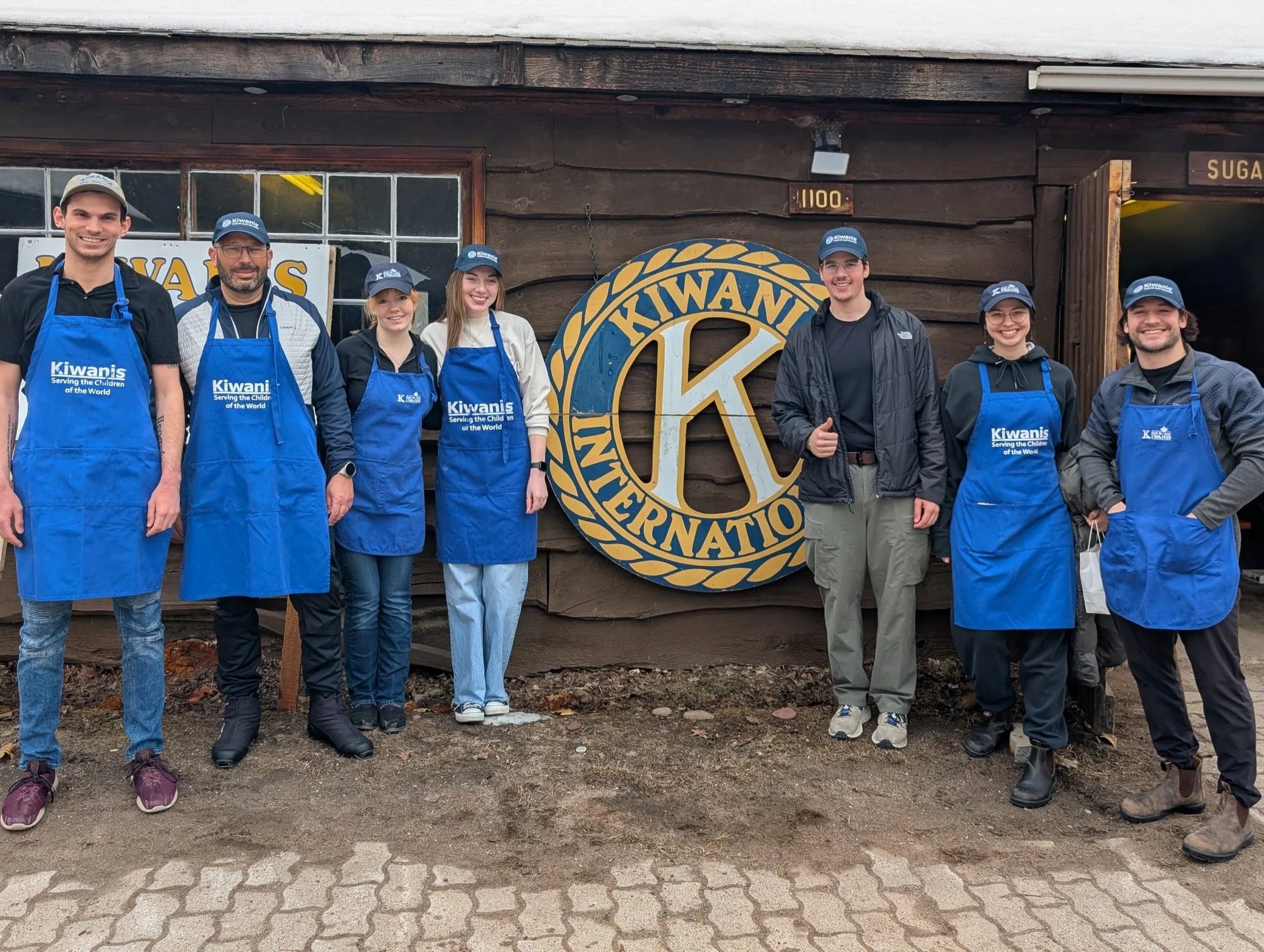 A group of seven people standing in front of a building with a large Kiwanis International sign. Some are wearing blue aprons with 'Kiwanis Serving the Children of the World' written on them. Others are wearing hats with the Kiwanis logo. The group is smiling and posing for the photo.