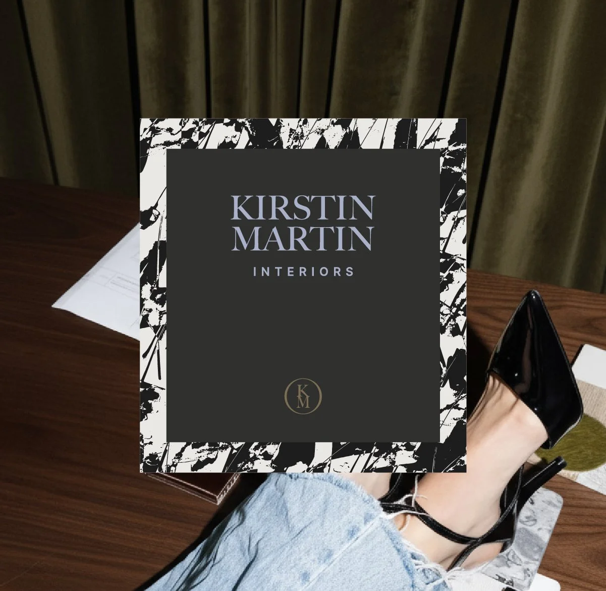 A sign reading 'Kirstin Martin Interiors' placed on a wooden table. A person's leg and foot in a black high heel shoe are visible, along with an open planner or notebook, a paper, and some clothing or fabric. The background has curtains.