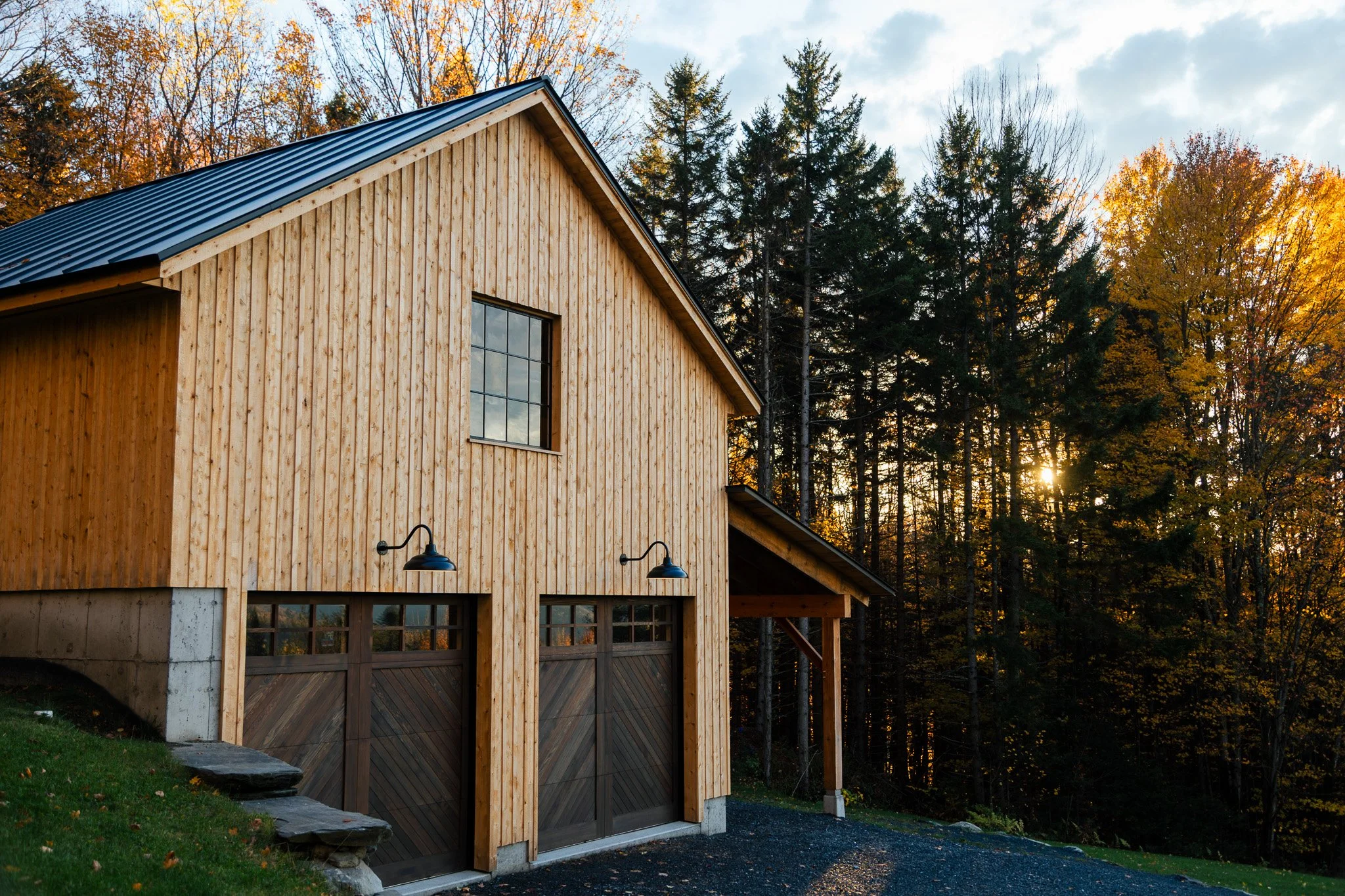 A two-story wooden house with a metal roof, garage doors, and outdoor lighting, set against a backdrop of autumn trees with the sun setting behind them.