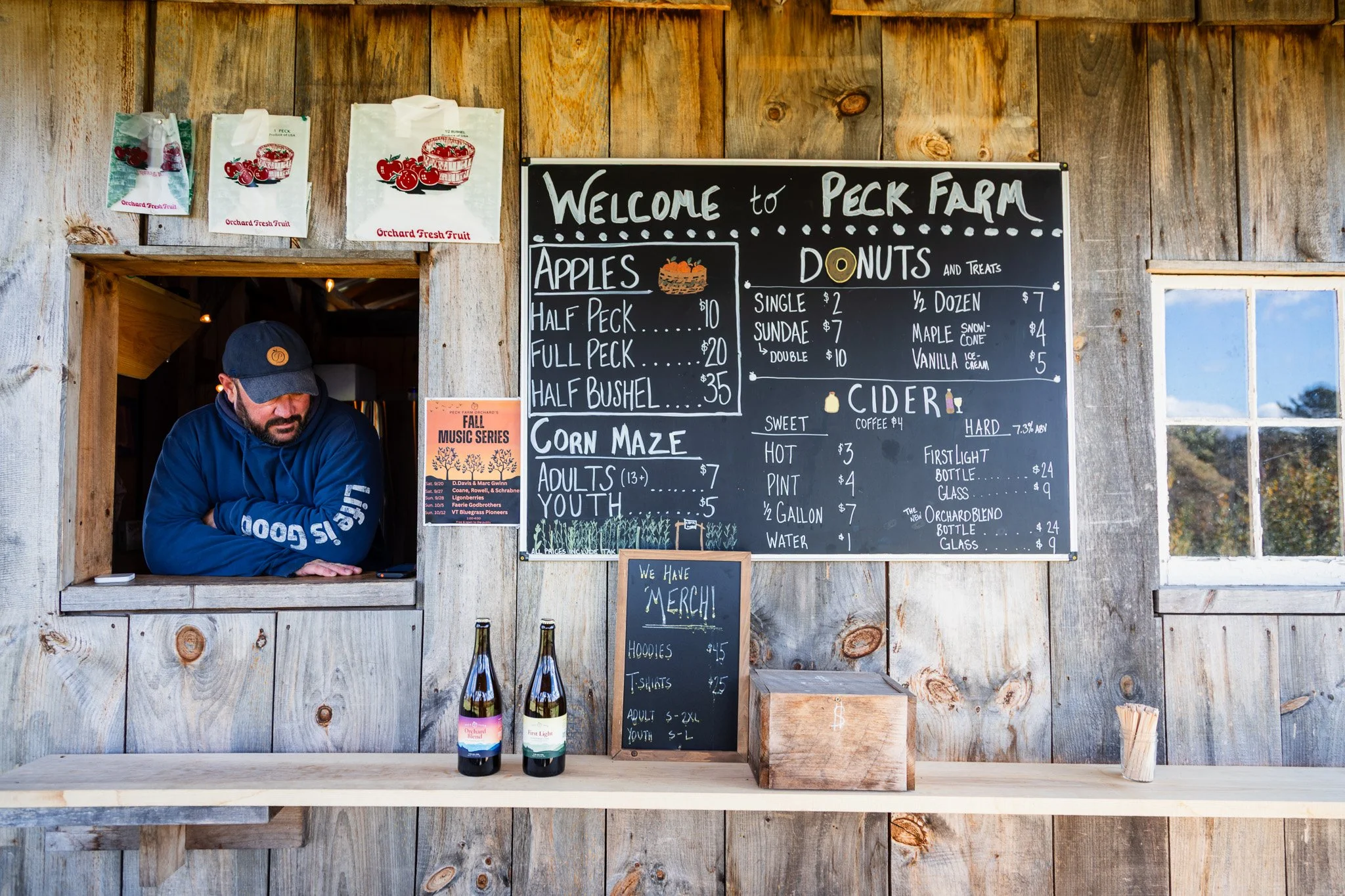 A man in a blue jacket and baseball cap leaning on a counter at Peck Farm, with a blackboard menu behind him listing apples, donuts, cider, and merchandise, along with bottles of drink on the counter.