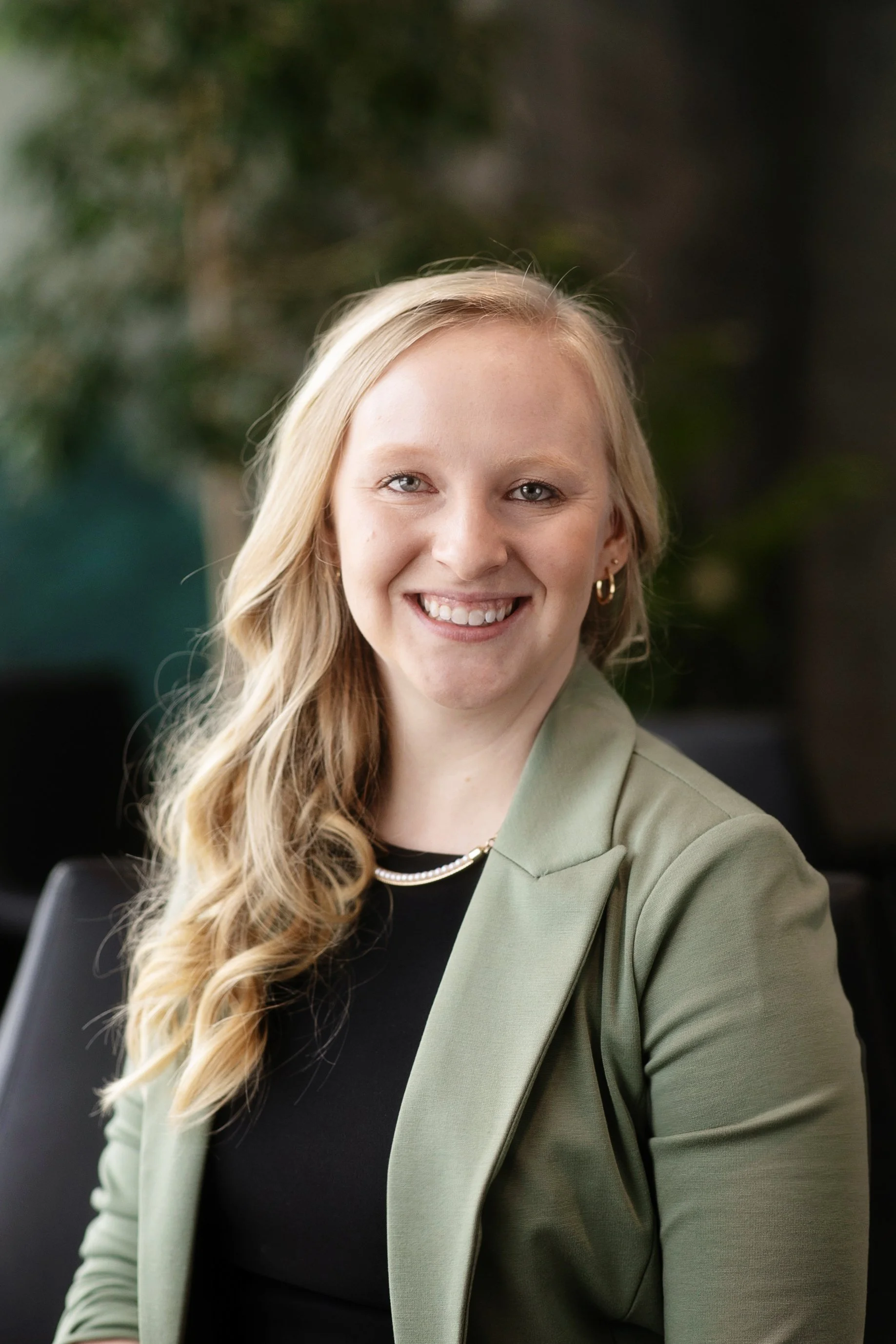 A young woman with long wavy blonde hair, smiling, wearing a light green blazer and gold hoop earrings, sitting in an indoor setting with a blurred green plant in the background.
