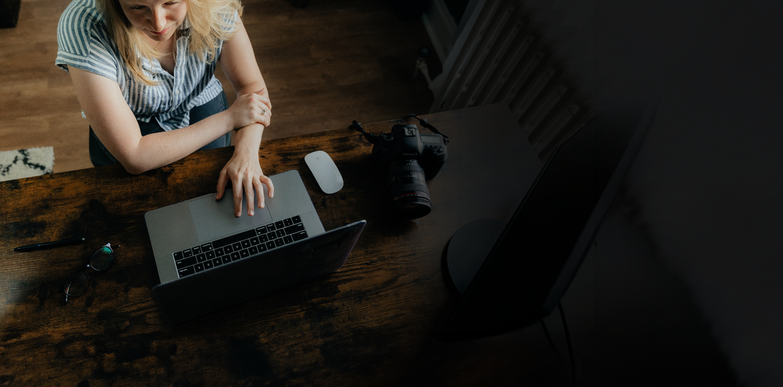 A woman sitting at a wooden desk using a laptop, with a camera, mouse, glasses, and pen nearby.