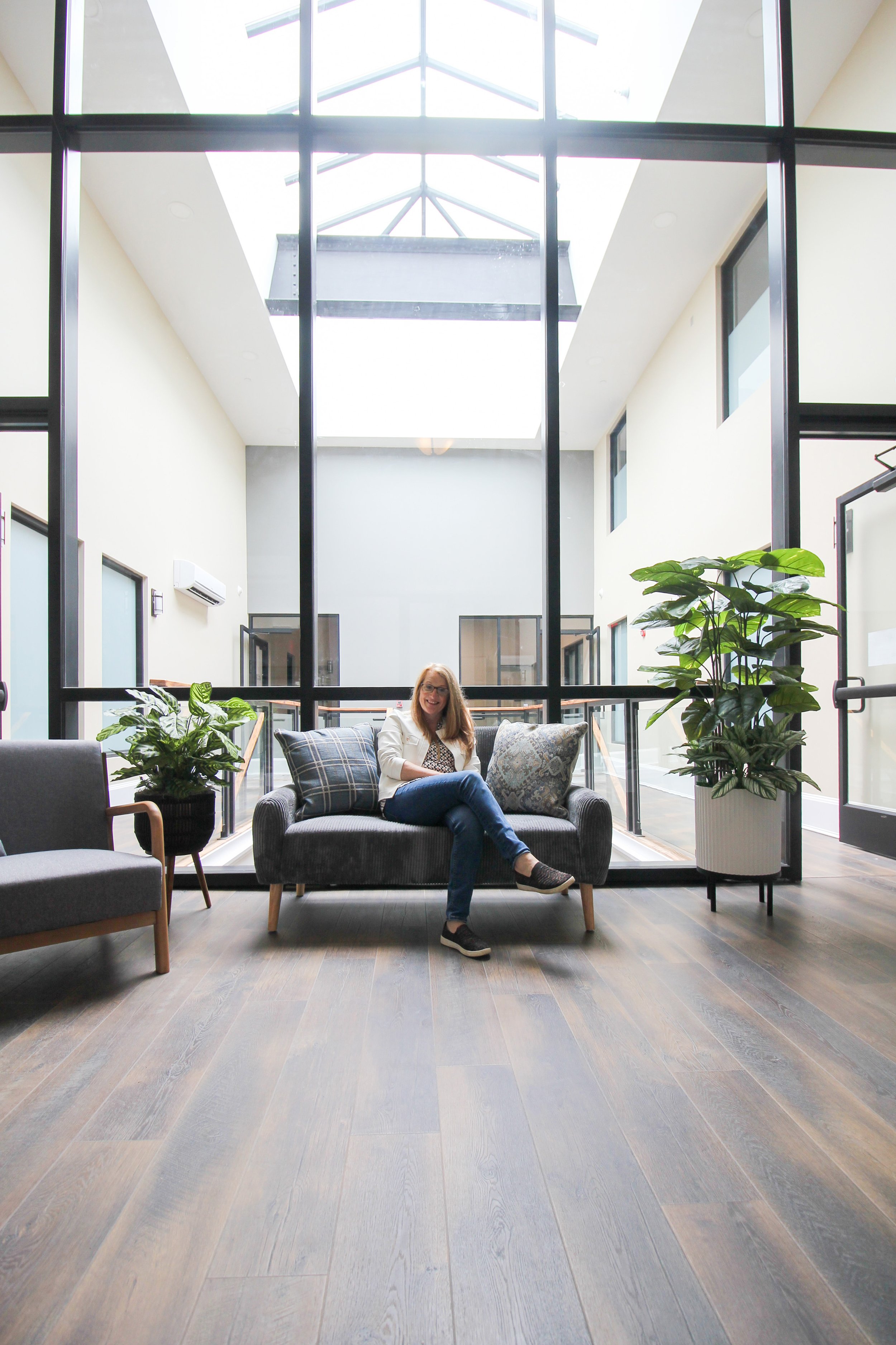 A woman sitting on a grey sofa in a modern indoor space with large windows, potted plants, and wooden flooring.