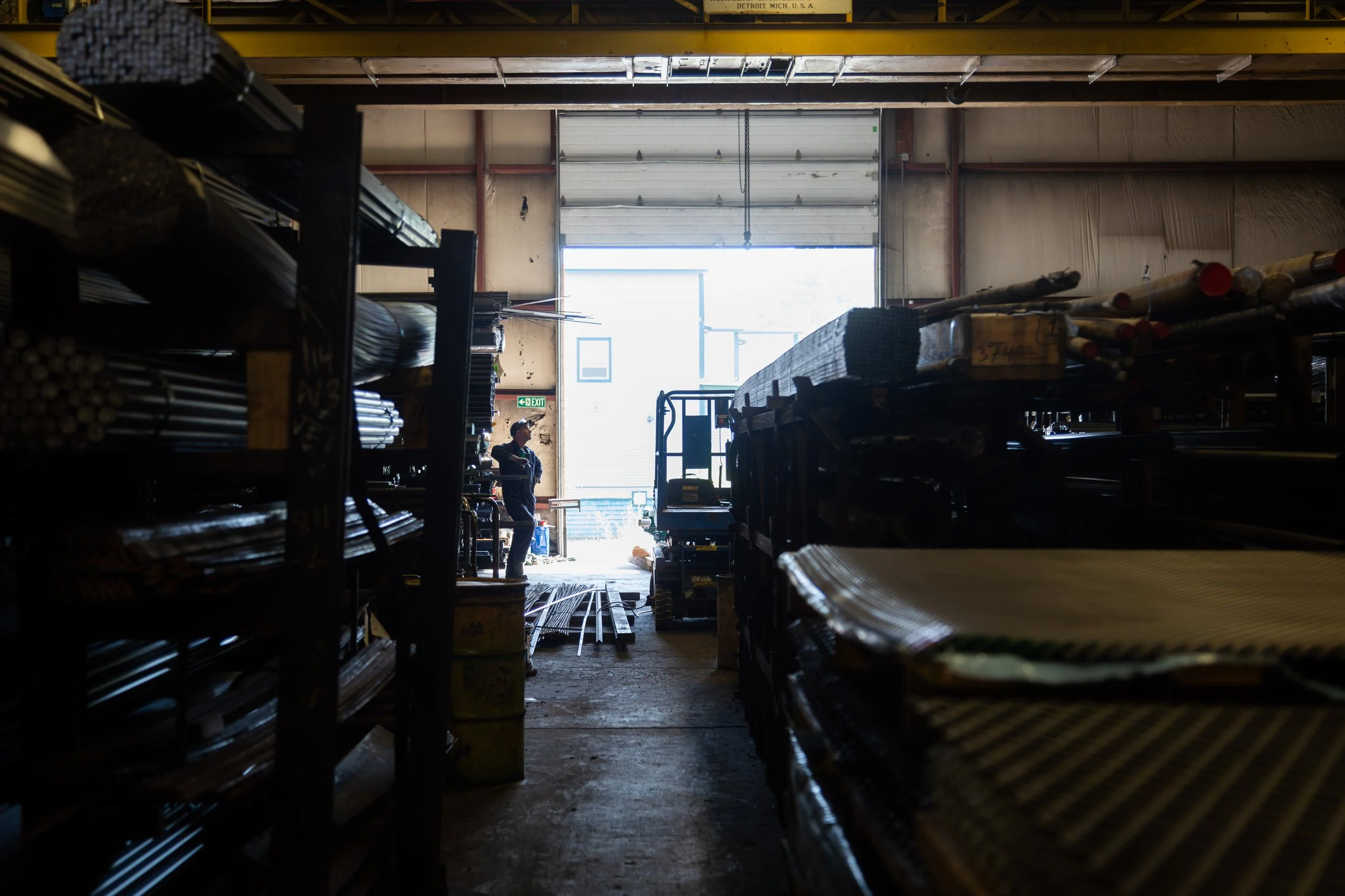 Silhouette of a person standing inside a warehouse near the open garage door, surrounded by racks of metal pipes and materials, with bright sunlight coming in from outside.