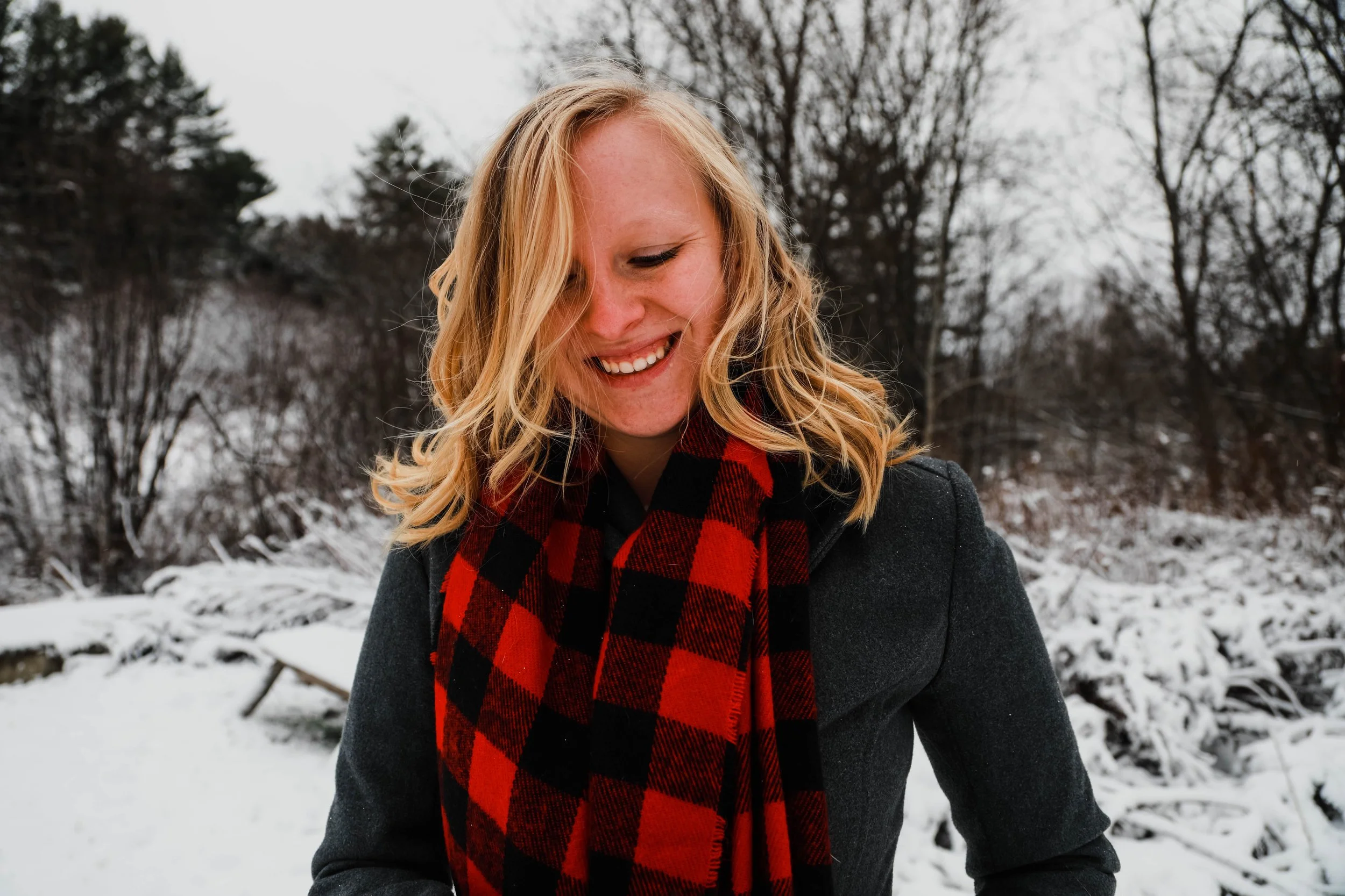 A young woman with blonde hair smiling and looking down, wearing a grey coat and a red and black checkered scarf, outdoors in a snowy landscape with trees in the background.