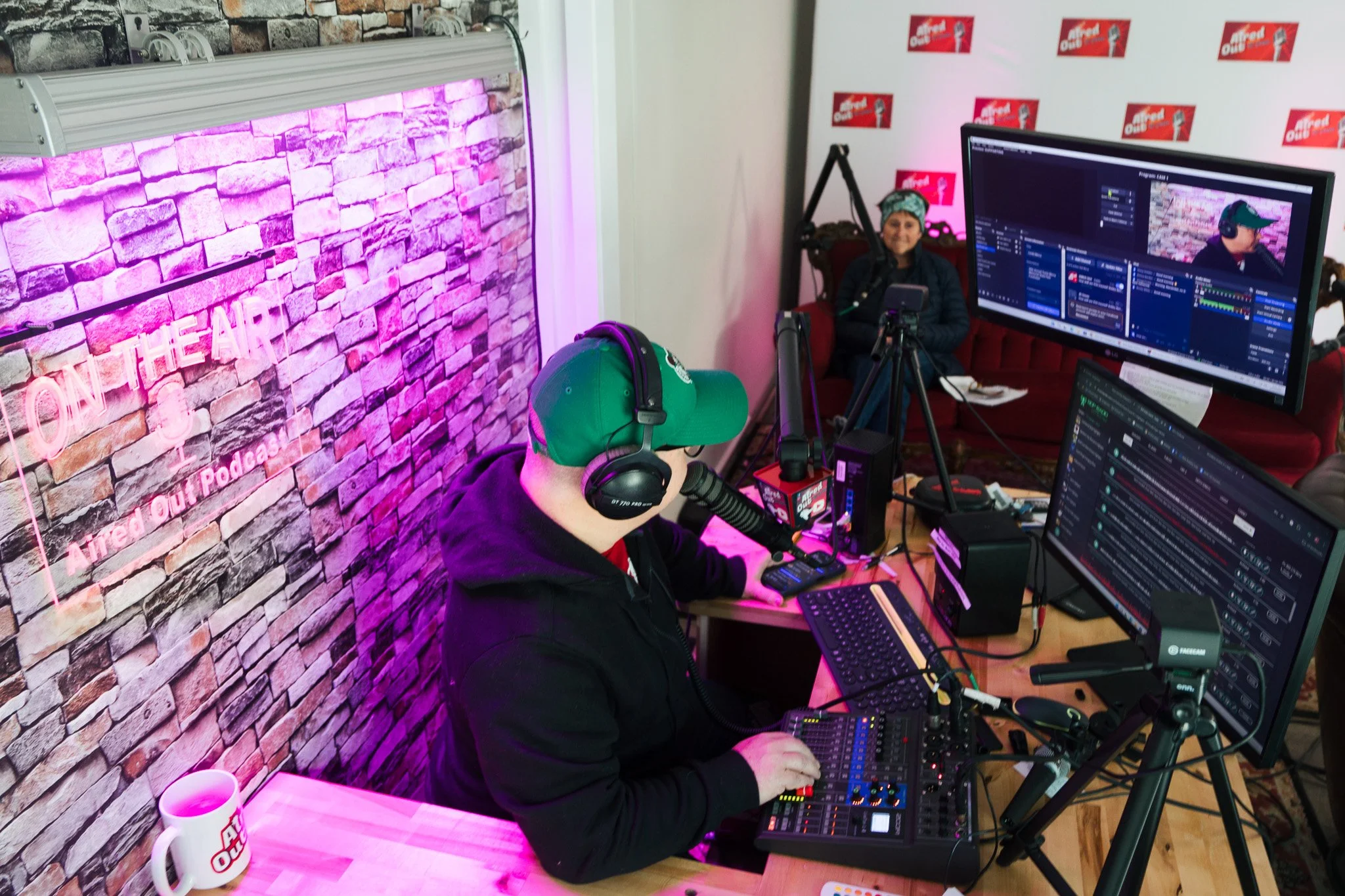 Two people in a podcast studio, one wearing headphones and a cap speaking into a microphone, with computers and sound equipment on the table, and a woman smiling in the background.