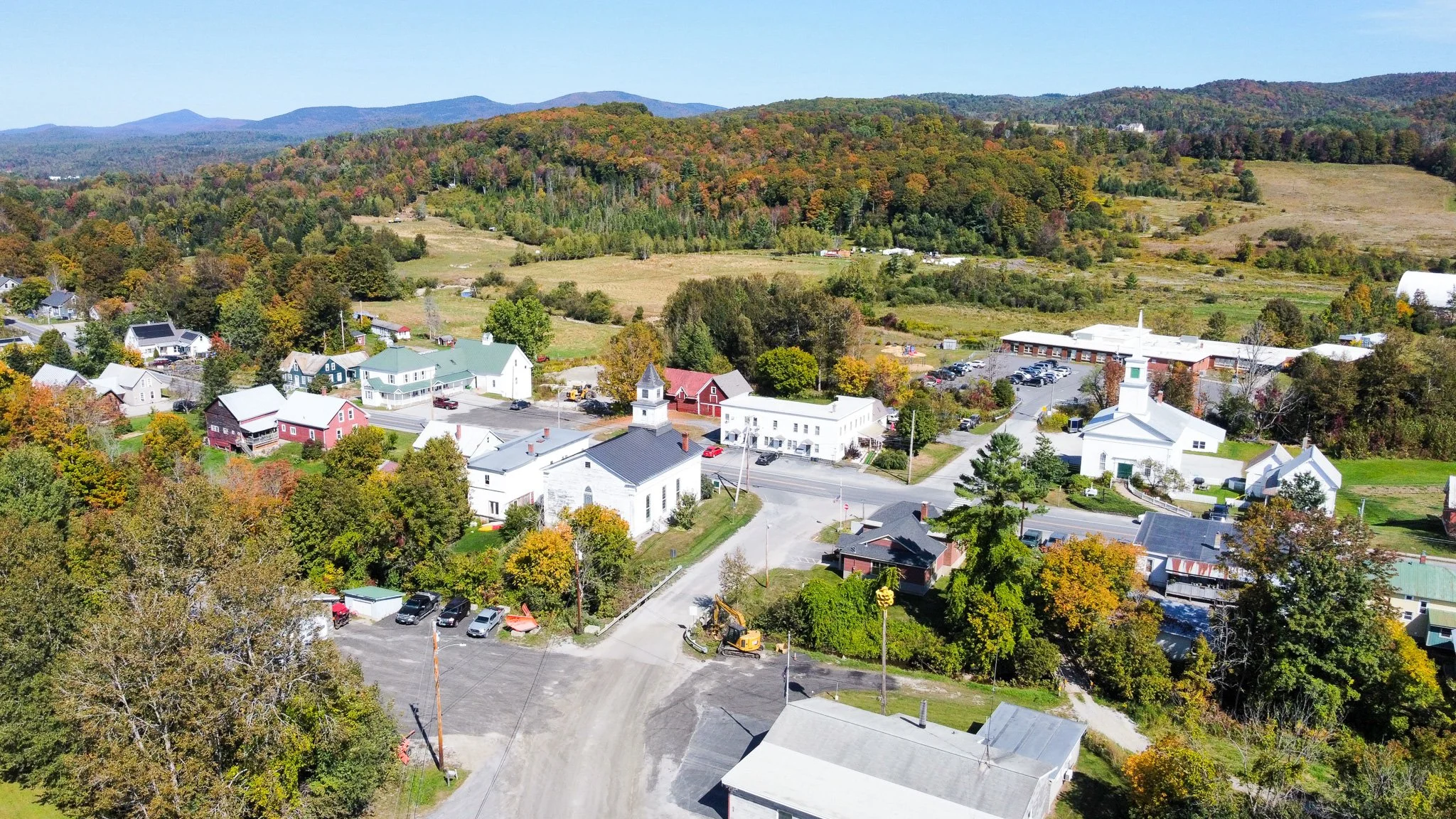 Aerial view of a small town with a mixture of white, red, and pastel-colored houses, a white church with a steeple, and a larger building with a parking lot. The town is surrounded by trees with fall foliage and open fields, with hills and mountains in the background on a sunny day.