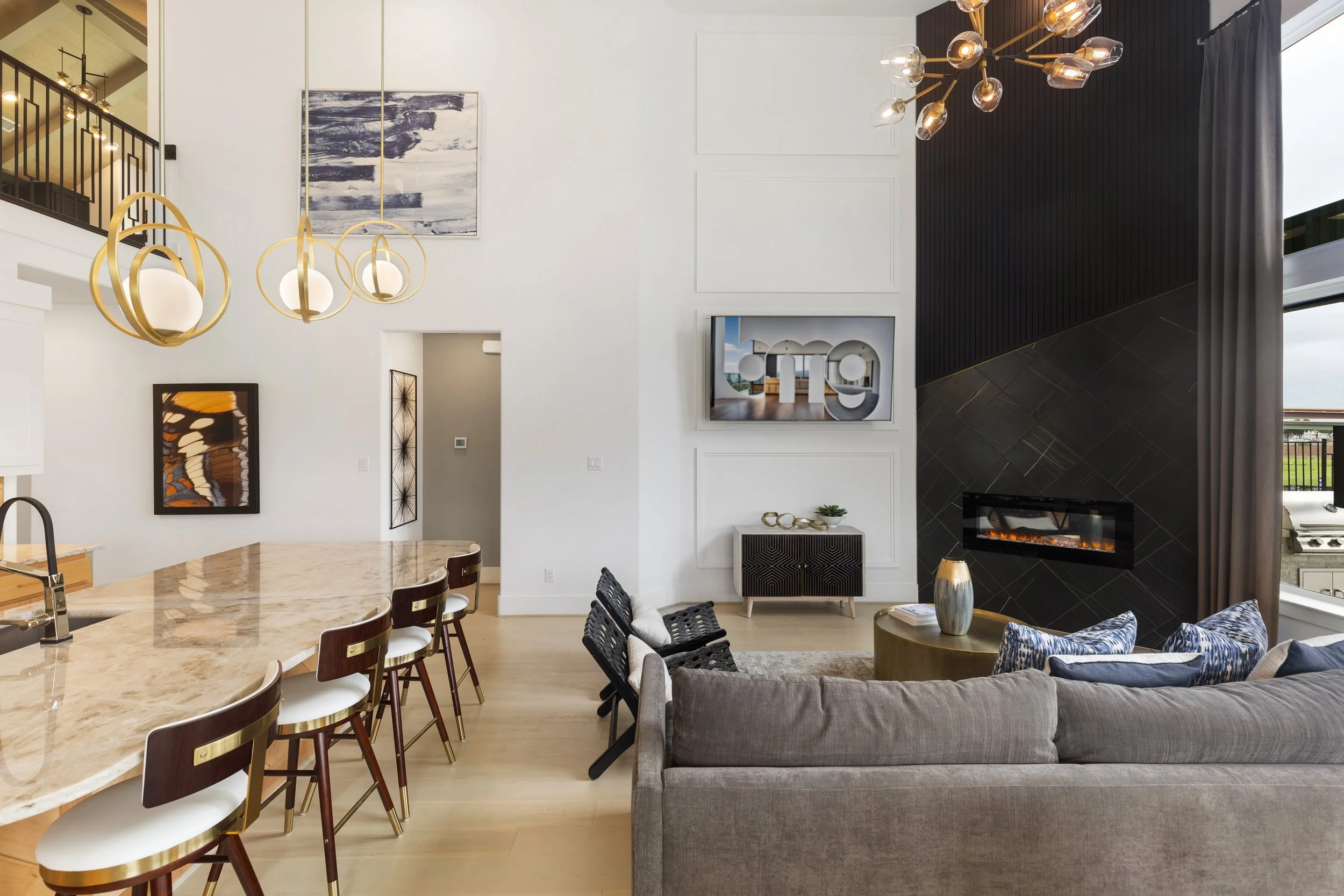 A modern open-concept living room and kitchen with high ceilings, white walls, abstract artwork, a black accent wall with a fireplace, a large gray sofa, a front-facing window with curtains, and a dining area with a marble-top island and wooden chair