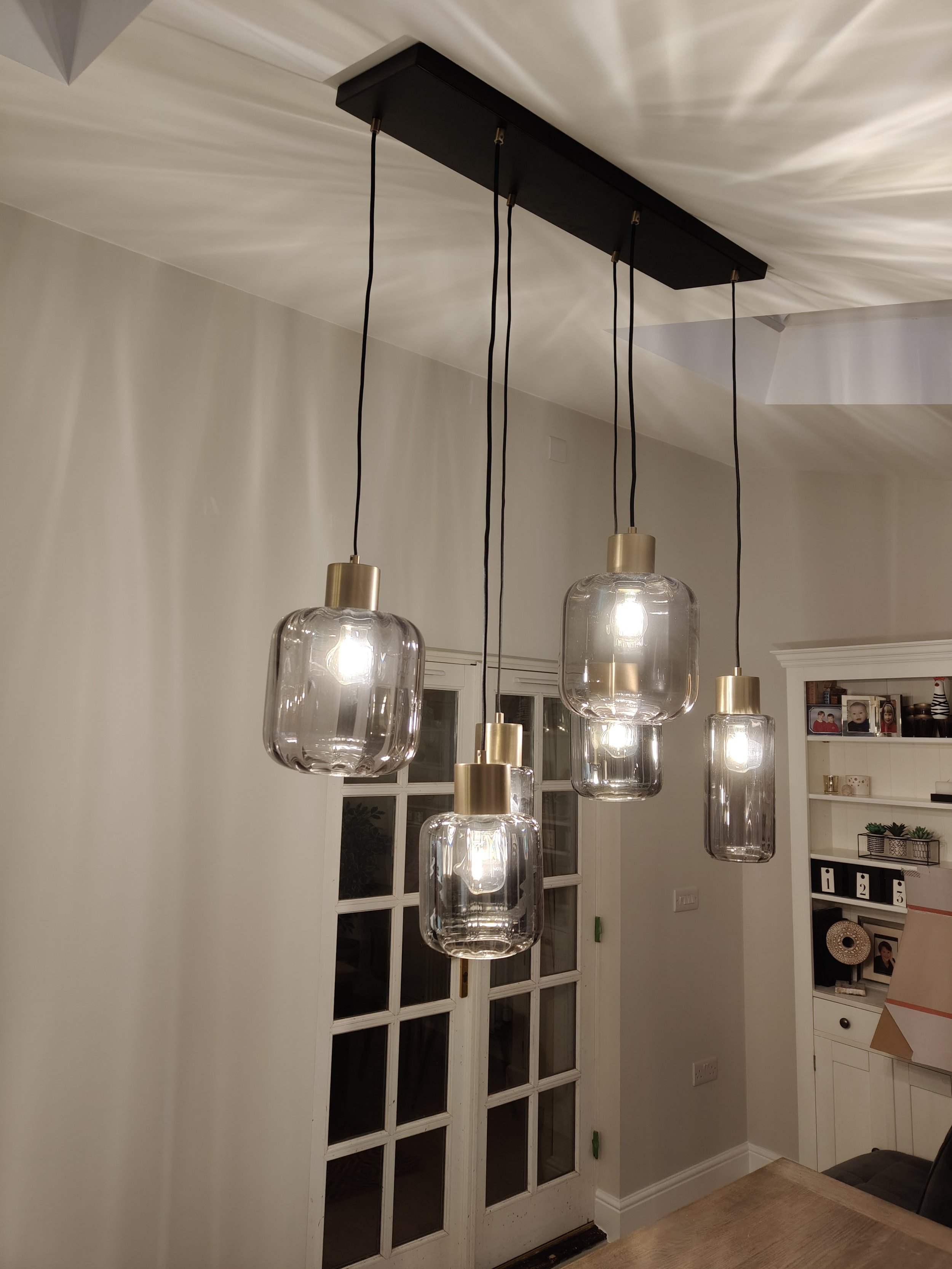 A modern ceiling light fixture with five glass pendant lights with gold fittings hanging over a wooden dining table in a bright room.