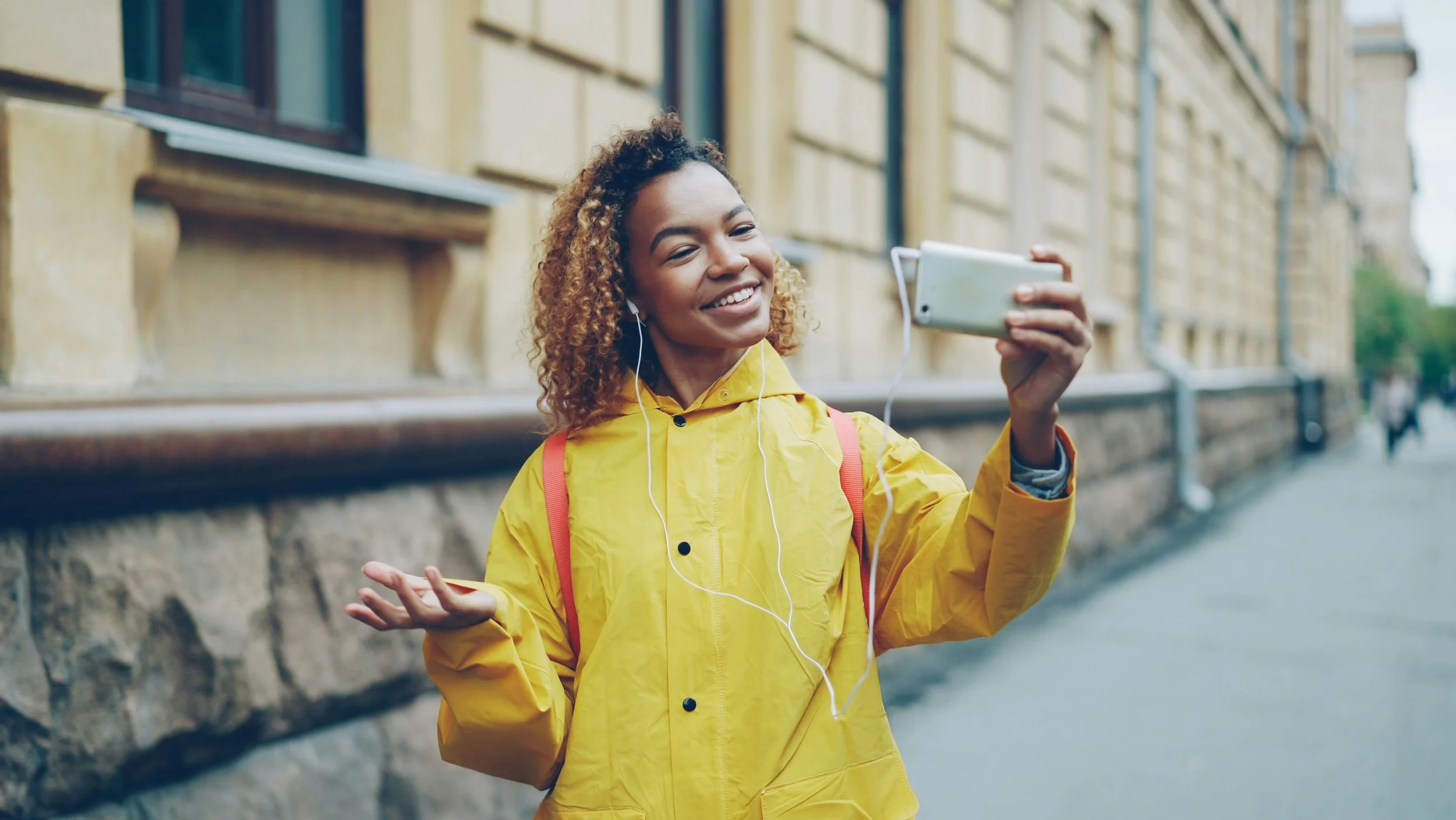 A young woman with curly hair, wearing a yellow rain jacket, is taking a selfie with her smartphone on a city sidewalk. She is smiling and has earphones in her ears.
