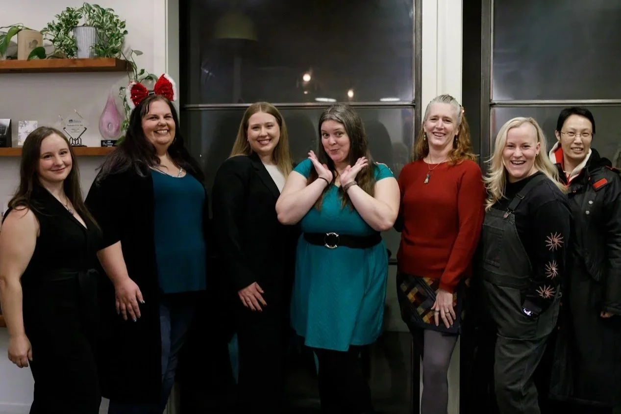 Group of seven women standing together indoors, smiling and posing for a photo, with a window and houseplants in the background.