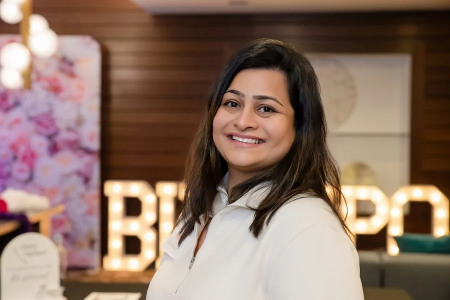 Smiling woman with dark hair wearing a white jacket indoors, with illuminated letters reading "biz expo" and floral decorations in the background.